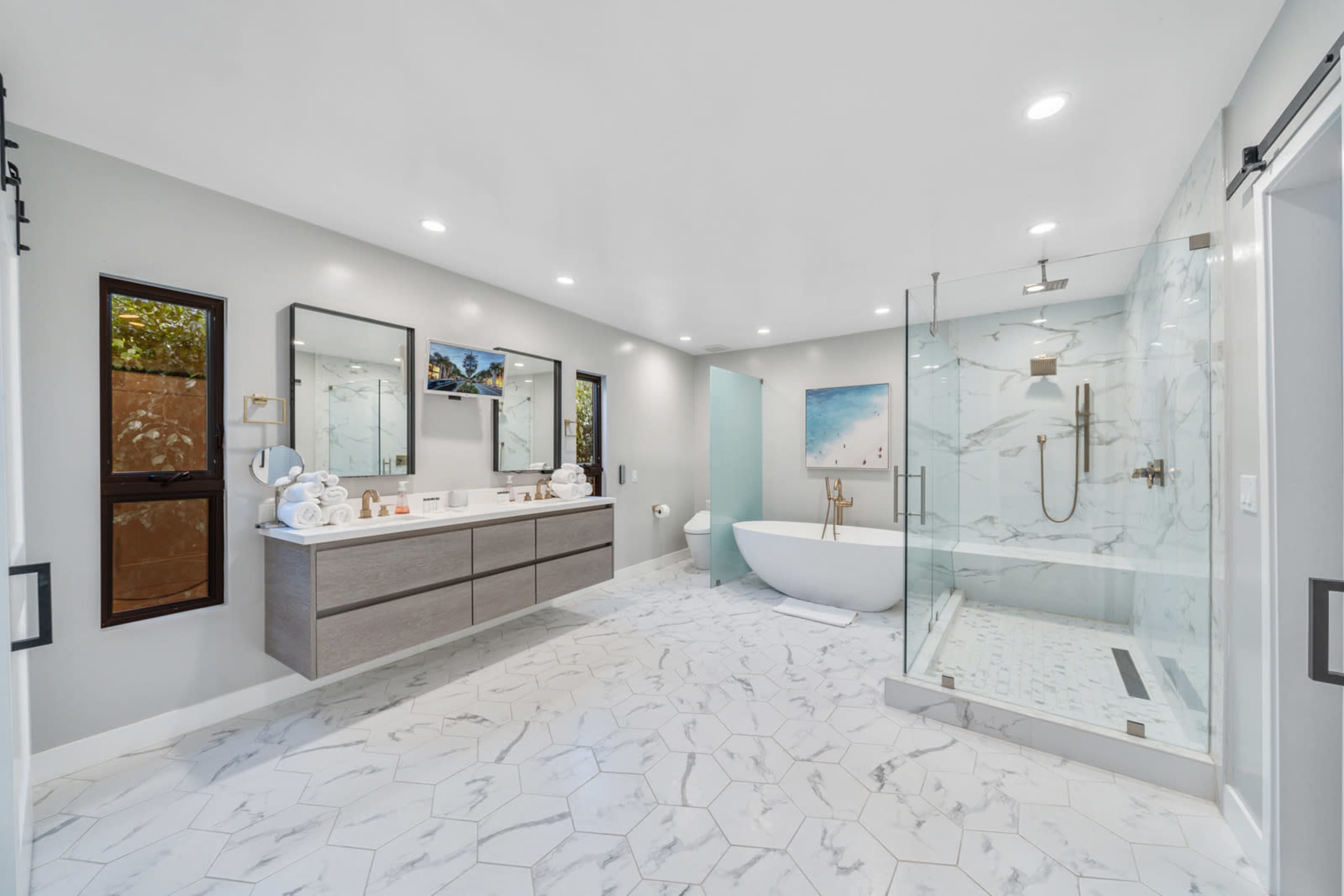 A modern bathroom features a freestanding bathtub, a glass-enclosed shower, dual vanities with mirrors, and a marble tile floor.