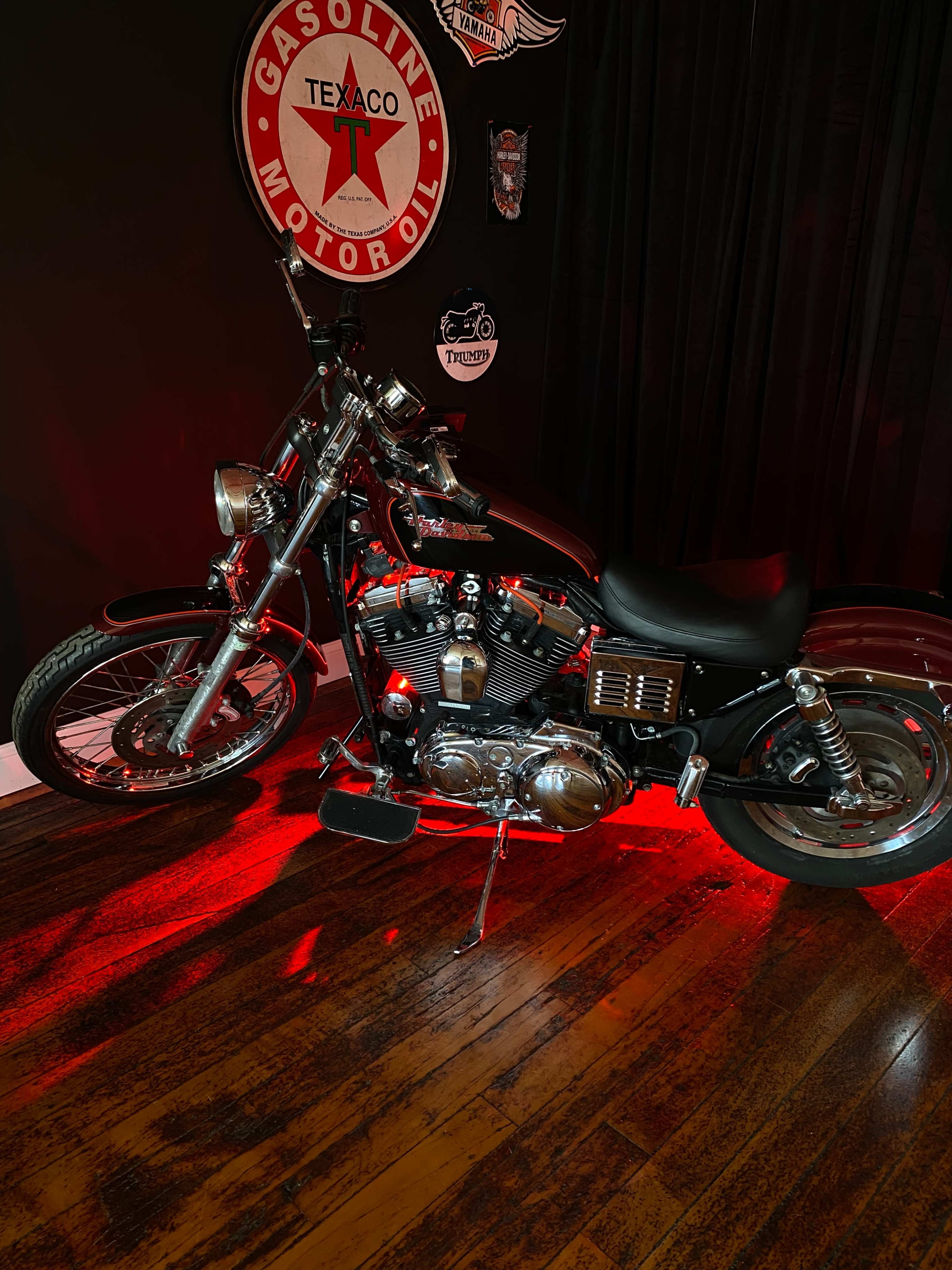 A motorcycle is parked on a wooden floor beneath red ambient lighting, with a vintage Texaco oil sign hanging on the wall behind it.