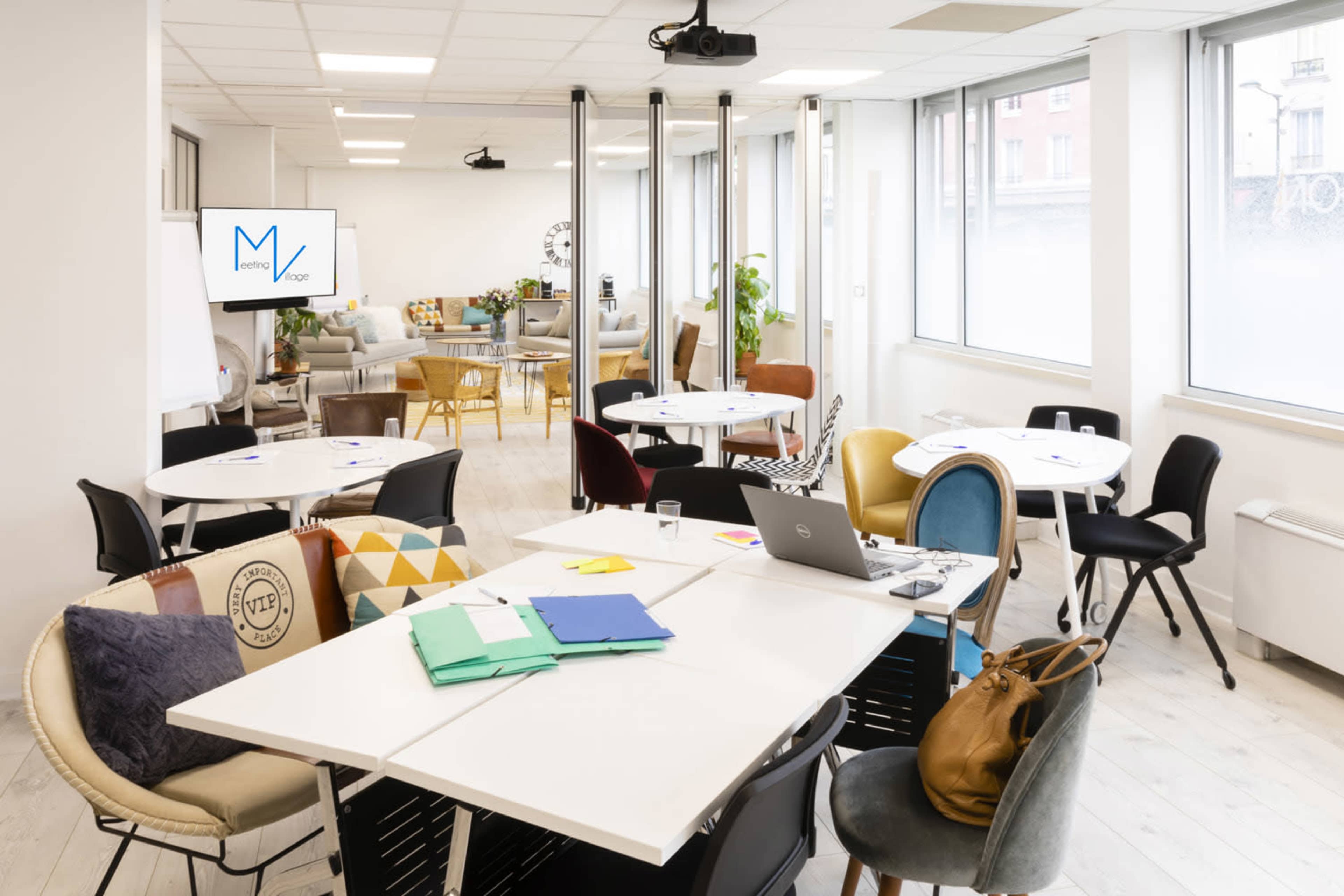 A modern, open-plan workspace featuring several tables and chairs, a projector, and windows allowing natural light.