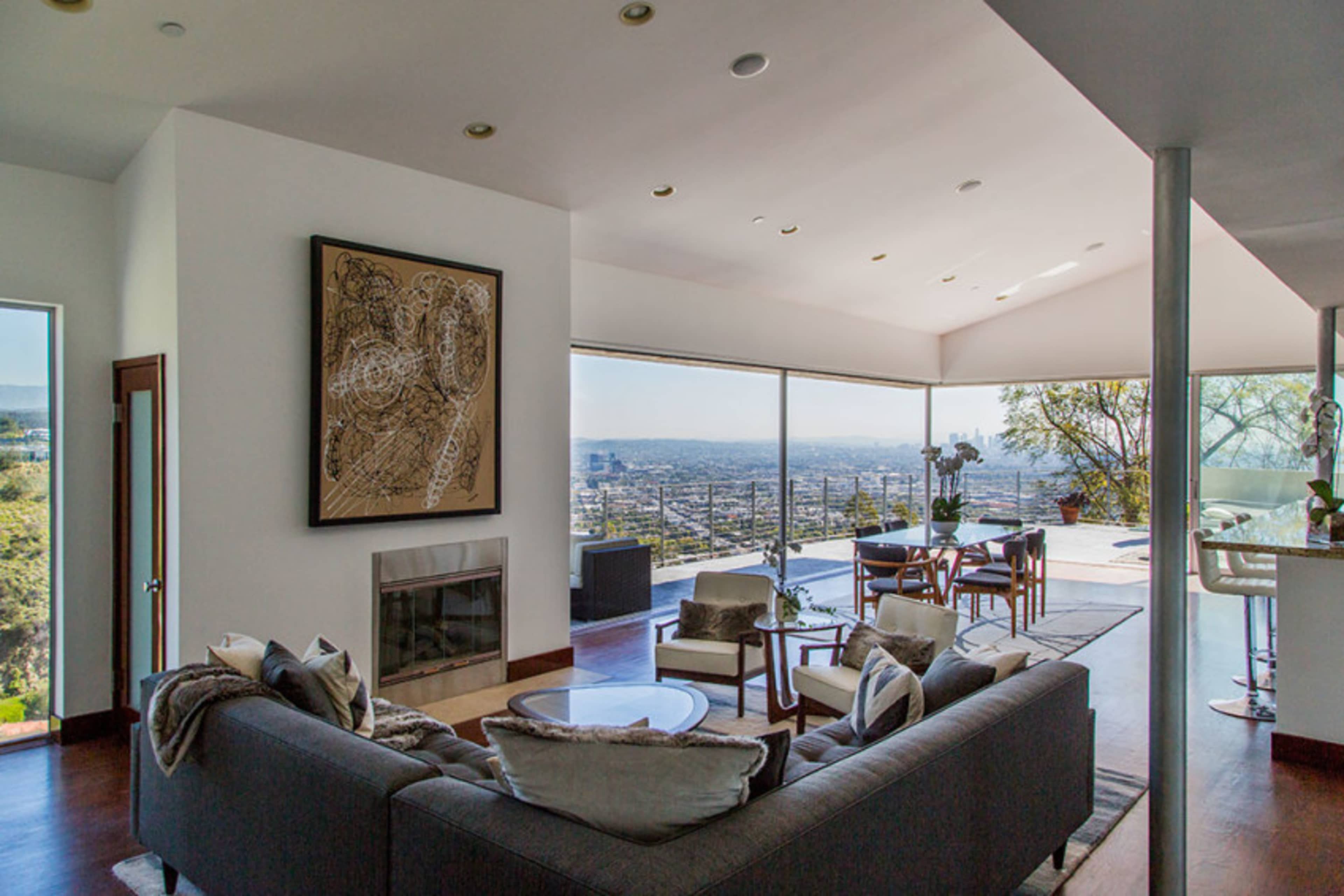 A modern living room with large windows offering a view of the city skyline, featuring a gray sofa, a glass coffee table, and a dining area in the background.