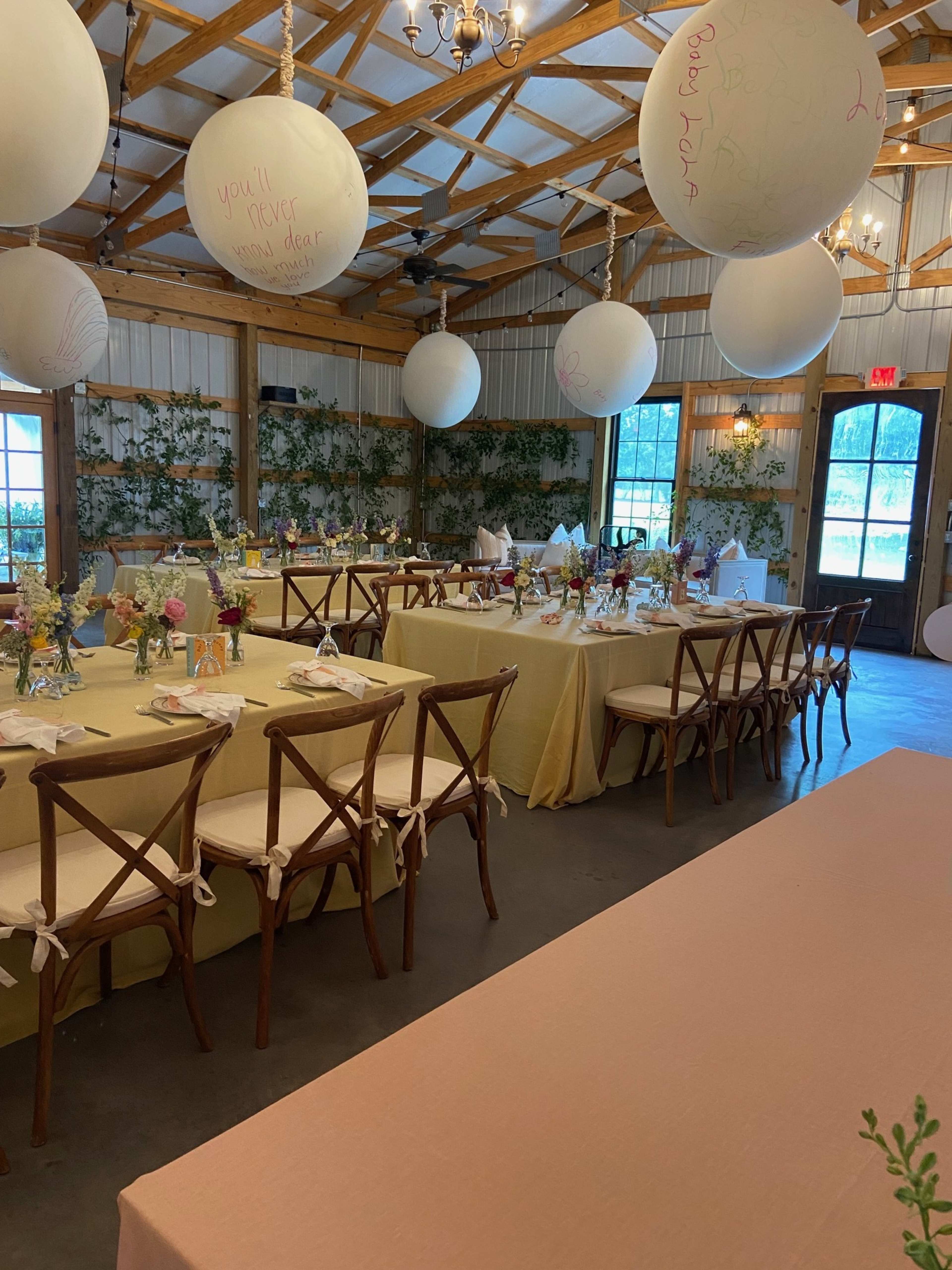 A reception hall features several tables covered with light-colored linens, floral centerpieces, and large balloons hanging above.