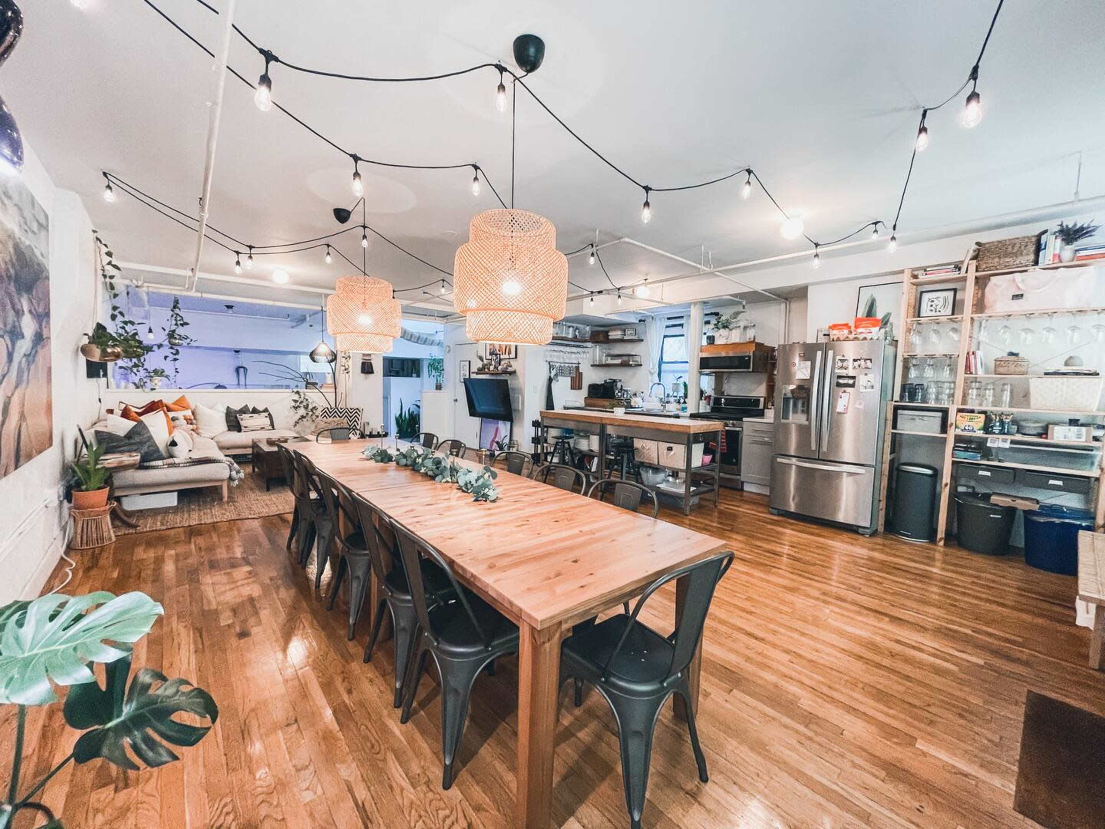 A spacious kitchen and dining area featuring a long wooden table surrounded by black chairs, overhead light fixtures, and a cozy seating arrangement.