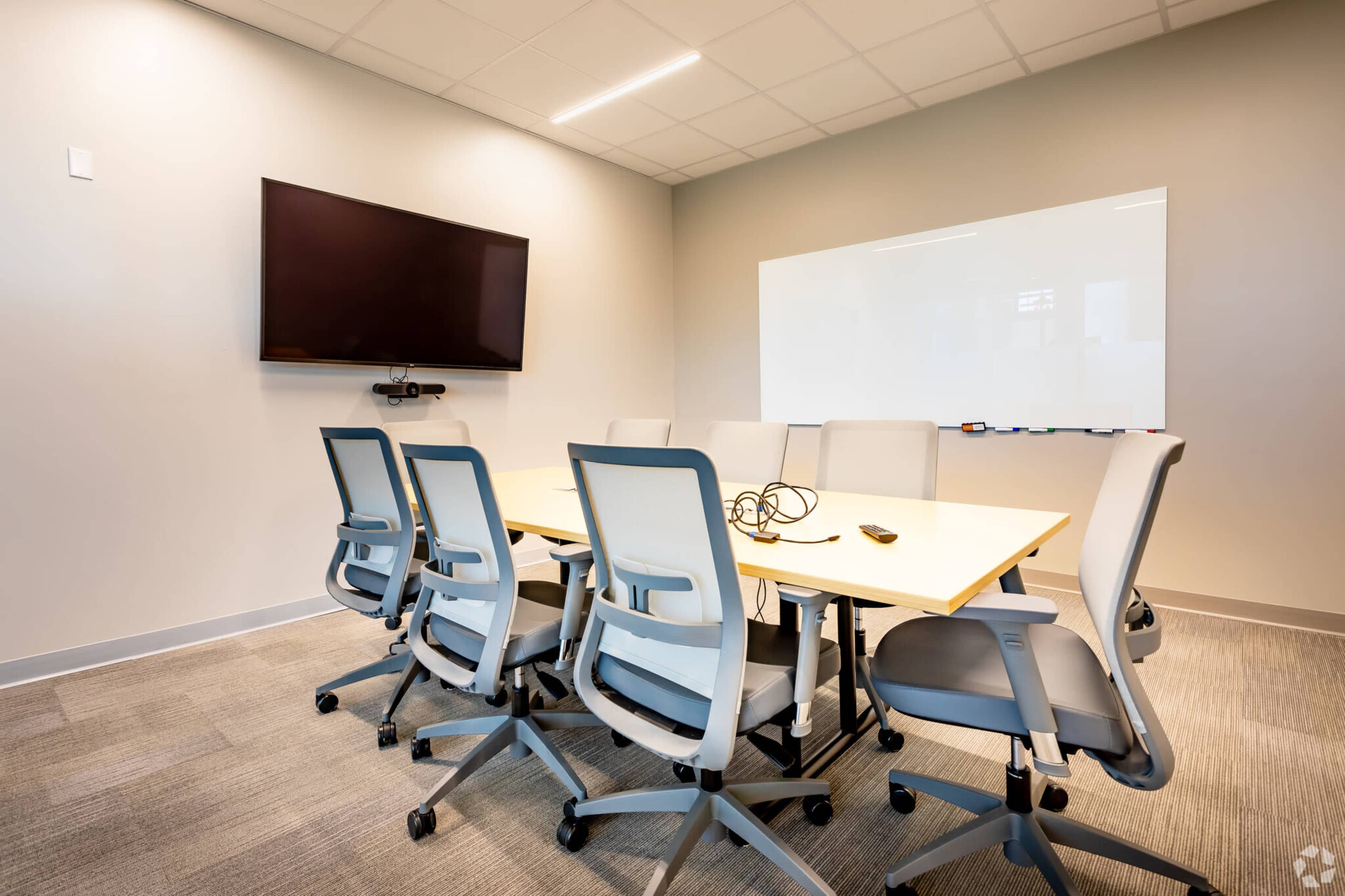 A modern conference room with a long table surrounded by six ergonomic chairs, a wall-mounted television, and a whiteboard.
