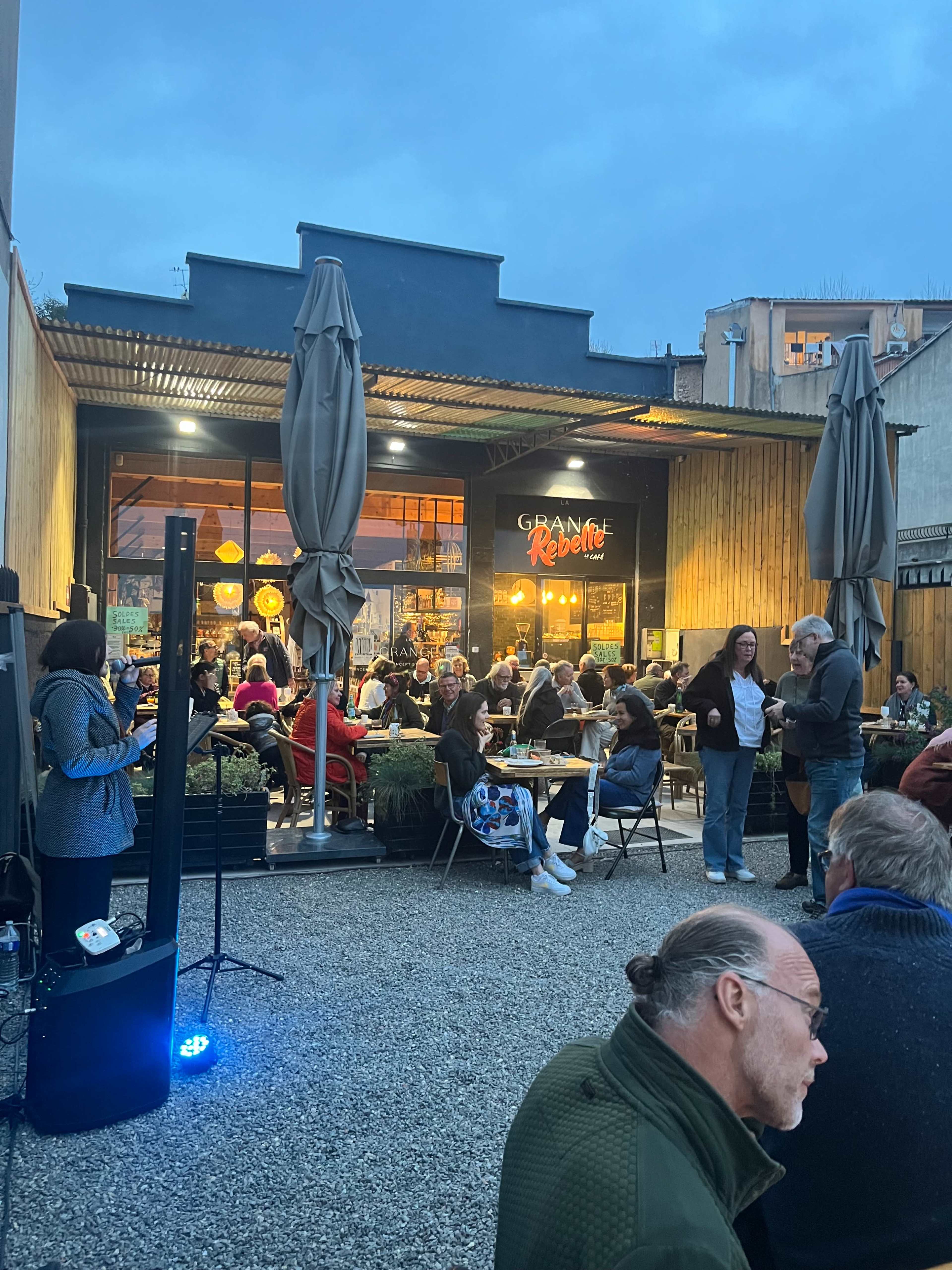 A bustling outdoor café scene features several patrons seated at tables, chatting and enjoying their evening as twilight sets in.