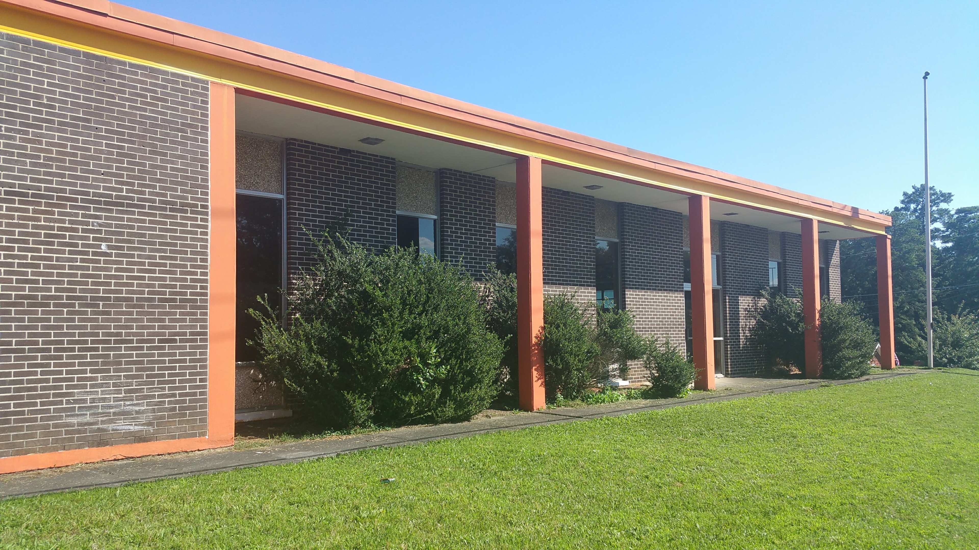 A one-story building with a brick facade and orange trim features several windows surrounded by greenery on a sunny day.