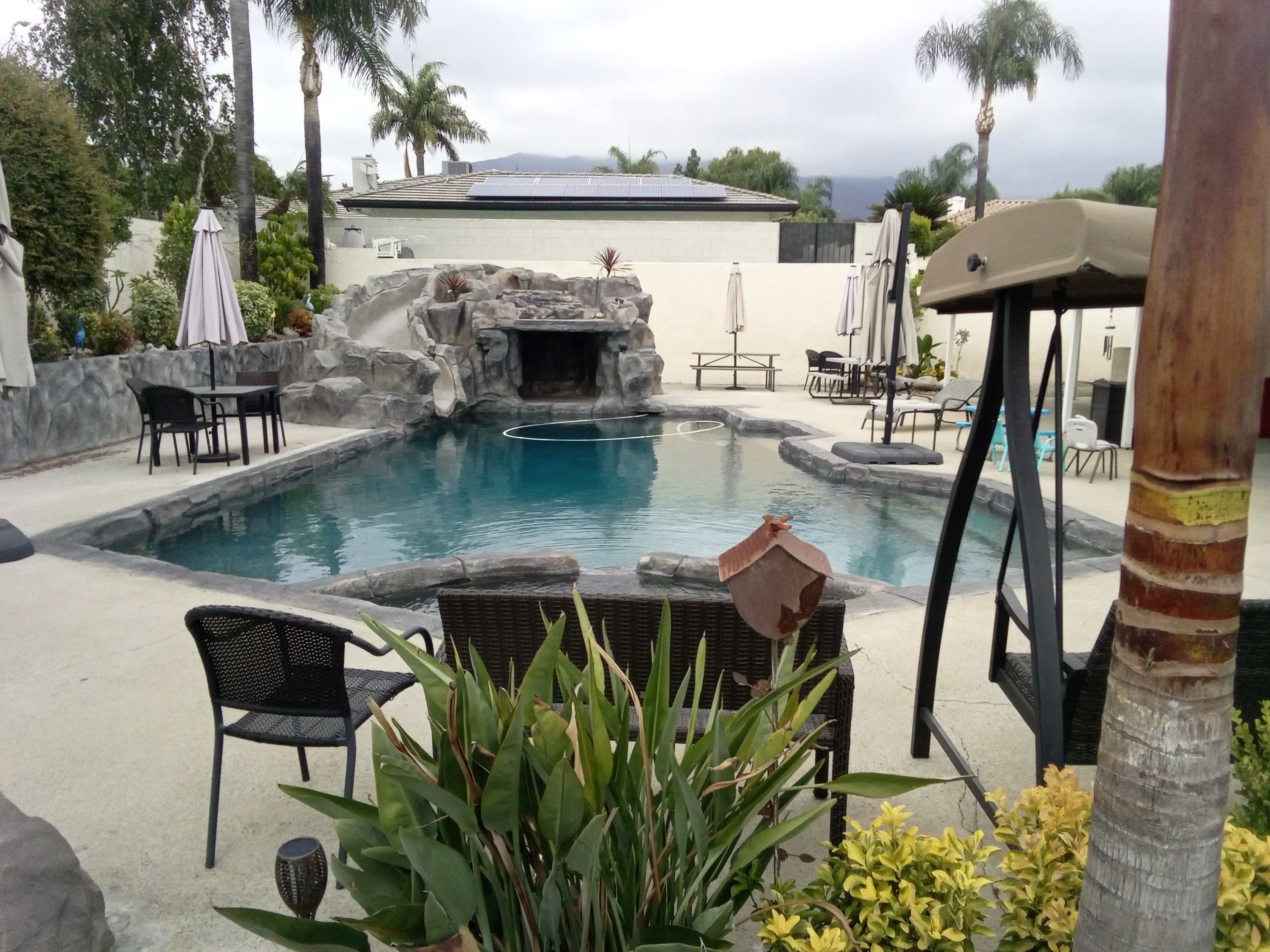 The image shows a landscaped pool area with a rock-feature design, surrounded by seating, tables, and palm trees under a cloudy sky.