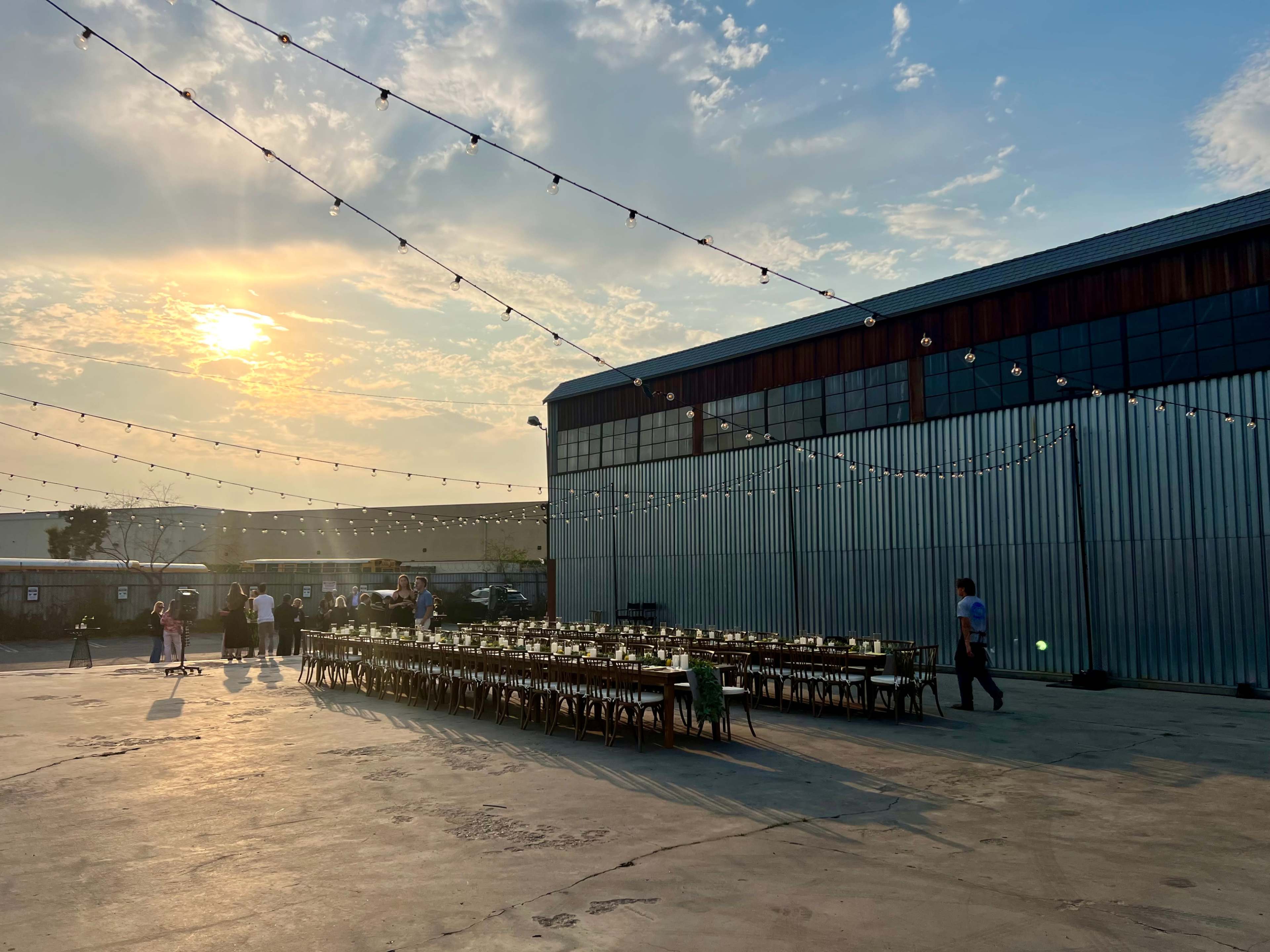 A long table with chairs is set up outside a warehouse, under string lights, as the sun sets in the background.