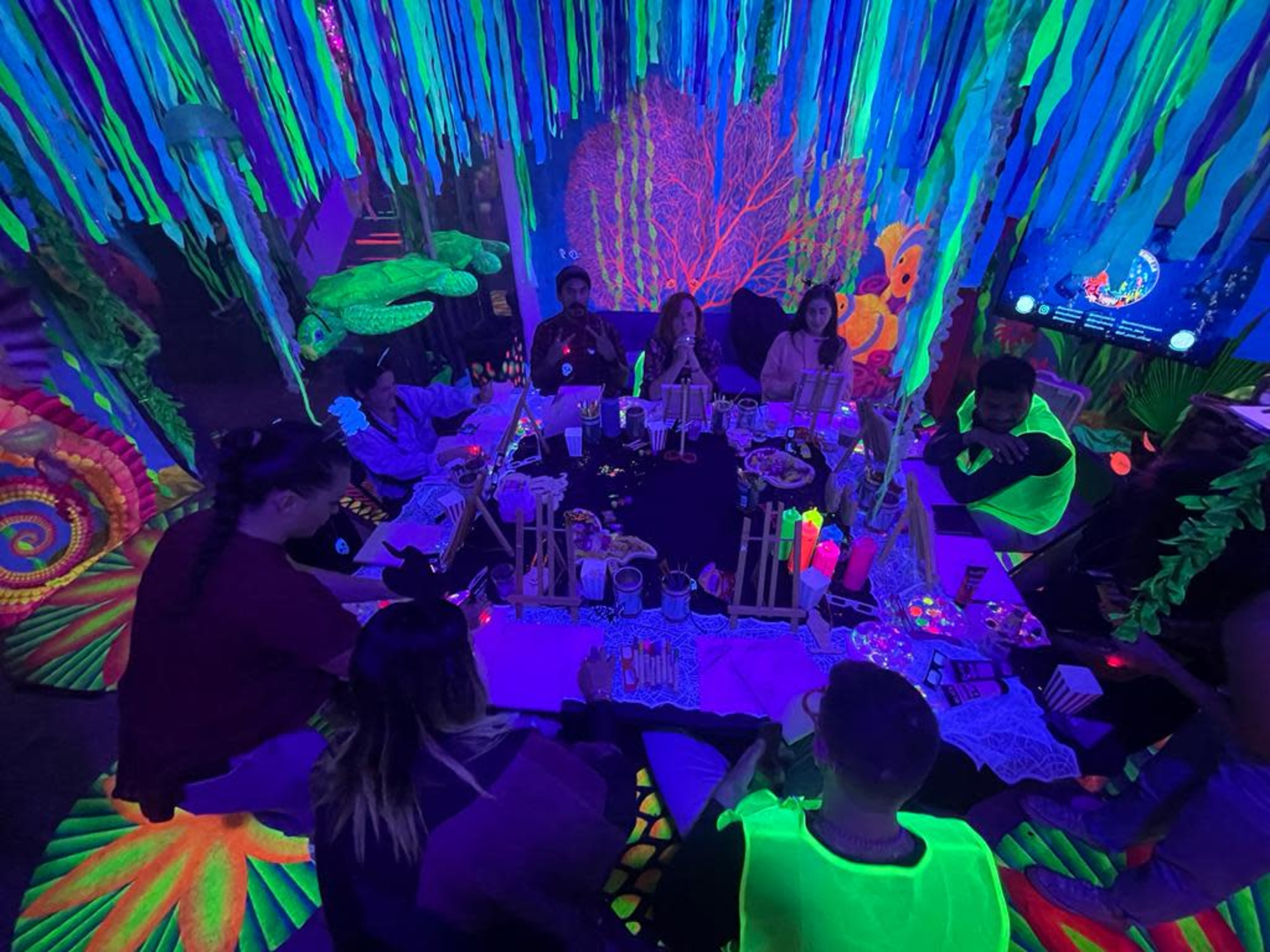 A group of people sits around a table in a brightly colored, neon-lit room decorated with glowing patterns and hanging decorations.