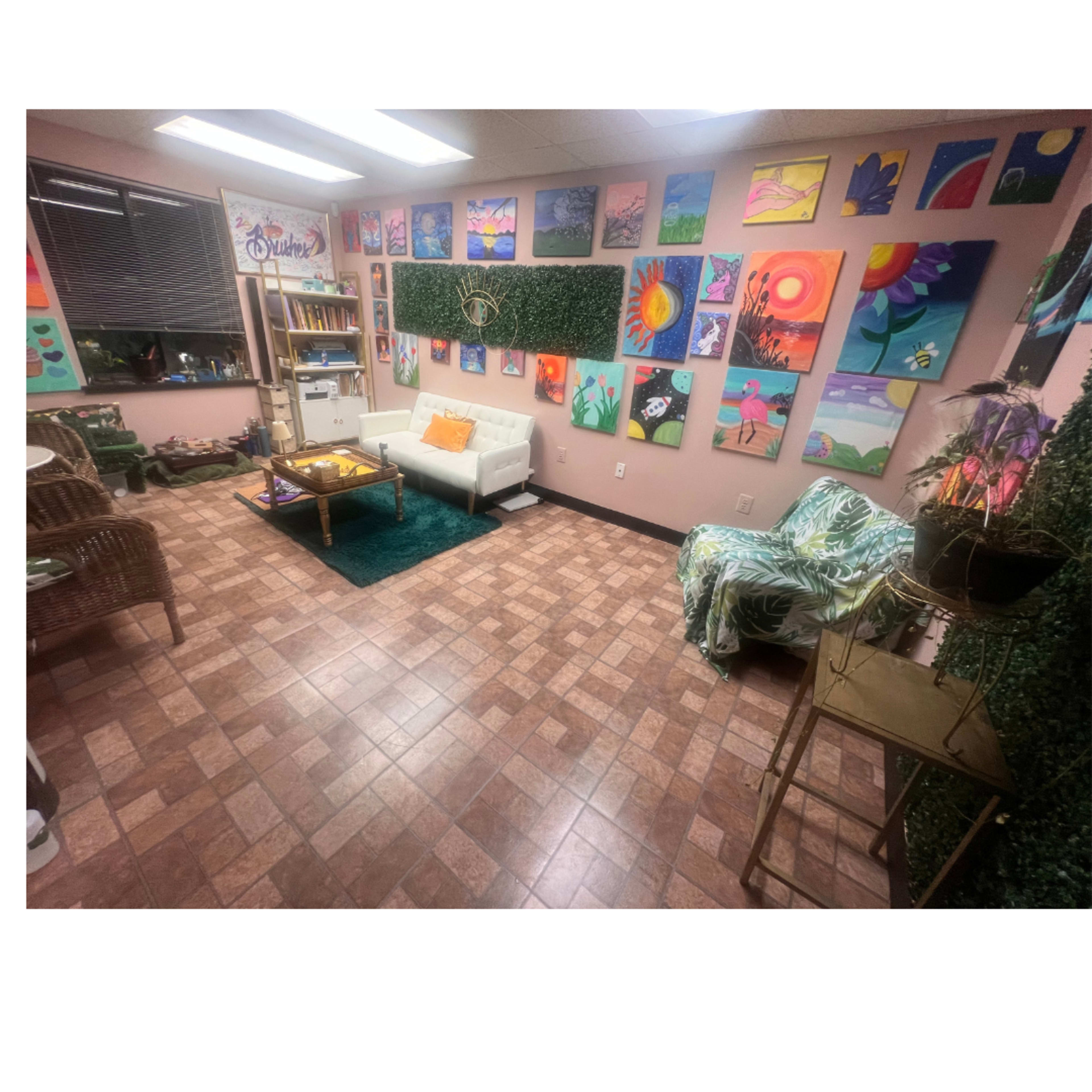 The image depicts a cozy room with a light brown tiled floor, featuring a white sofa, a coffee table, and a variety of colorful paintings displayed on the walls.