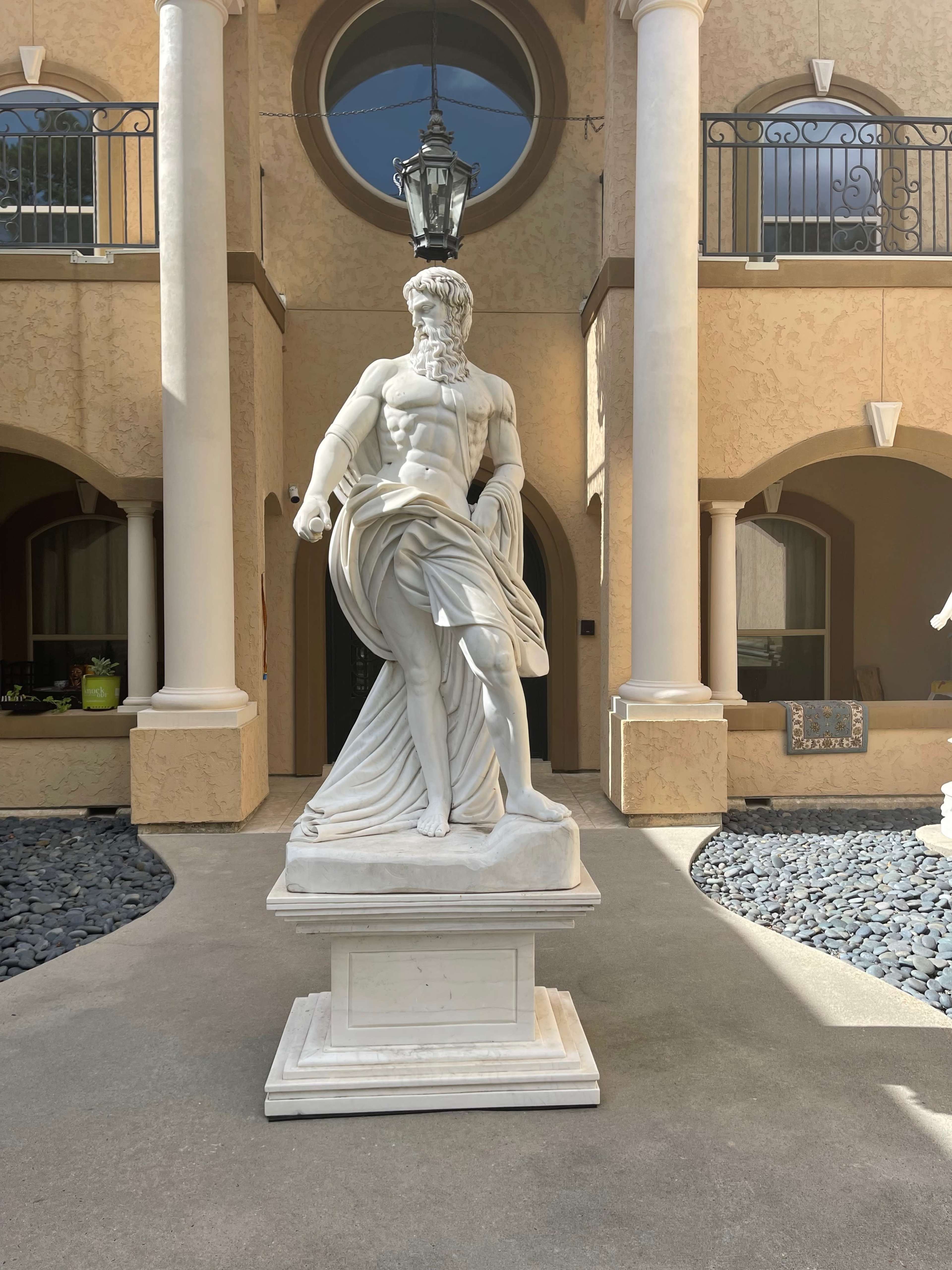 A tall white marble statue of a bearded figure draped in flowing garments, positioned in a courtyard with decorative columns and a lantern above.