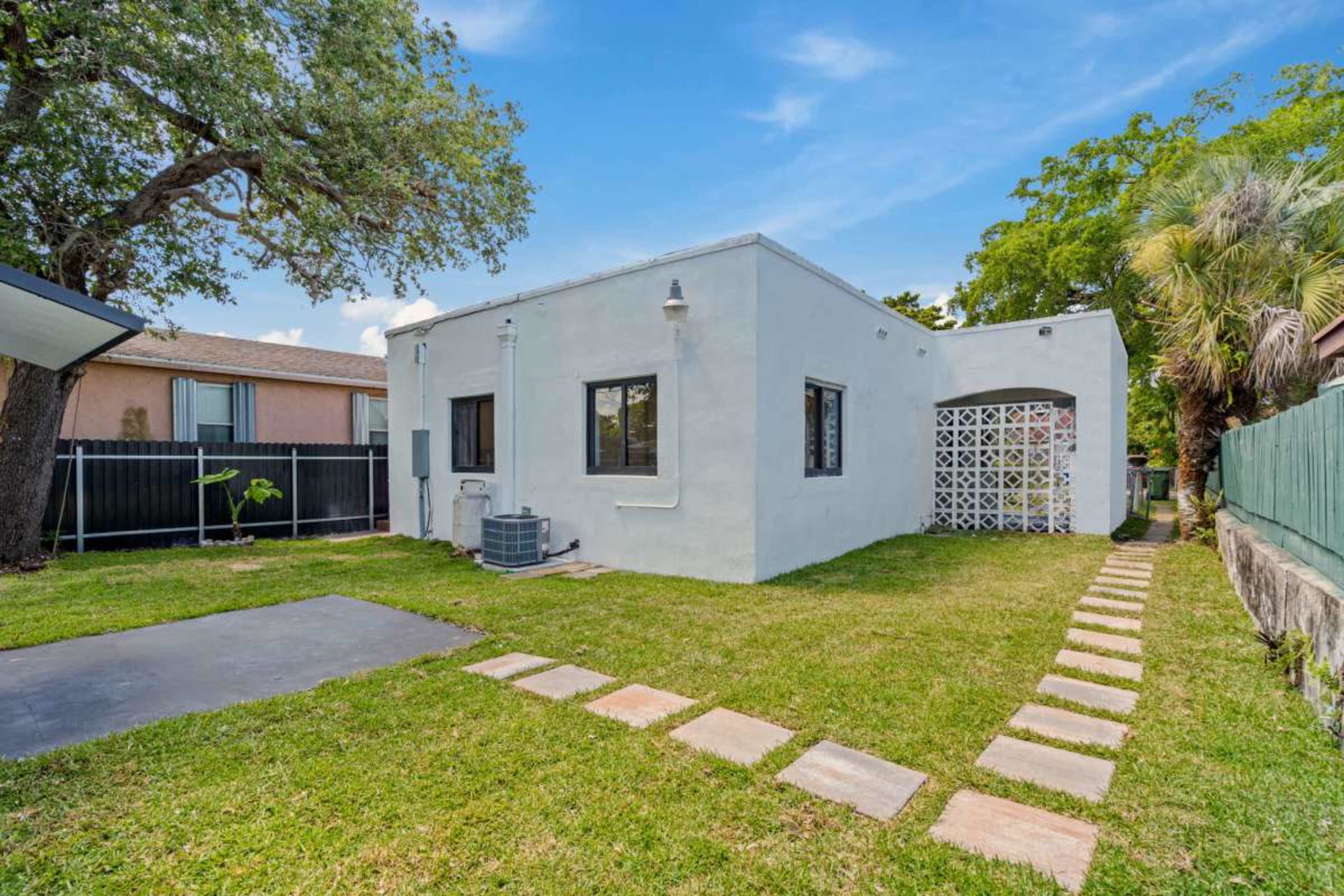 The image shows a white stucco house with a small patio area and a stone pathway leading to the entrance, surrounded by grass and trees.