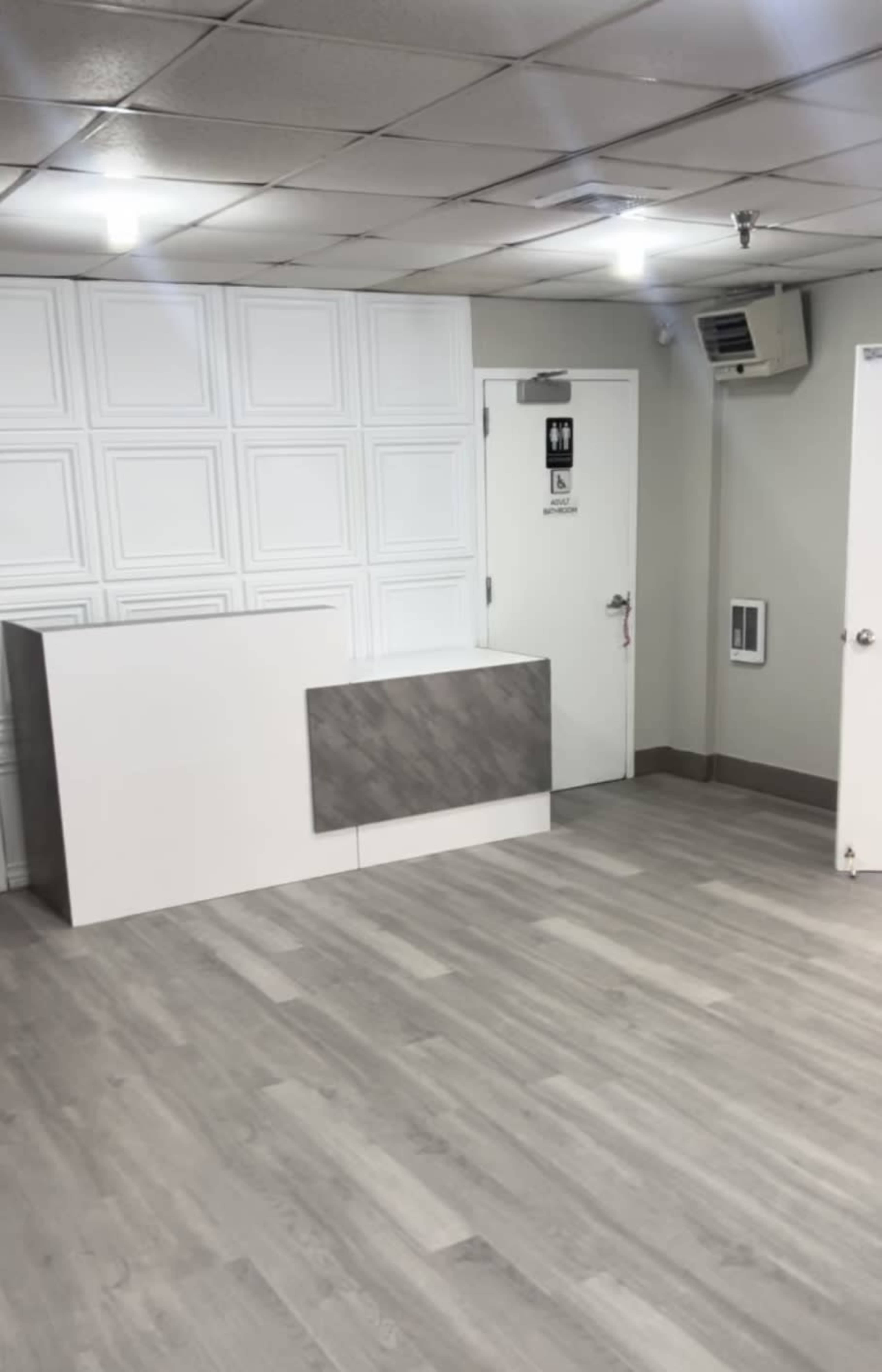 A minimalist interior space features a white reception desk against a paneled wall, with a restroom door and an air conditioning unit visible.