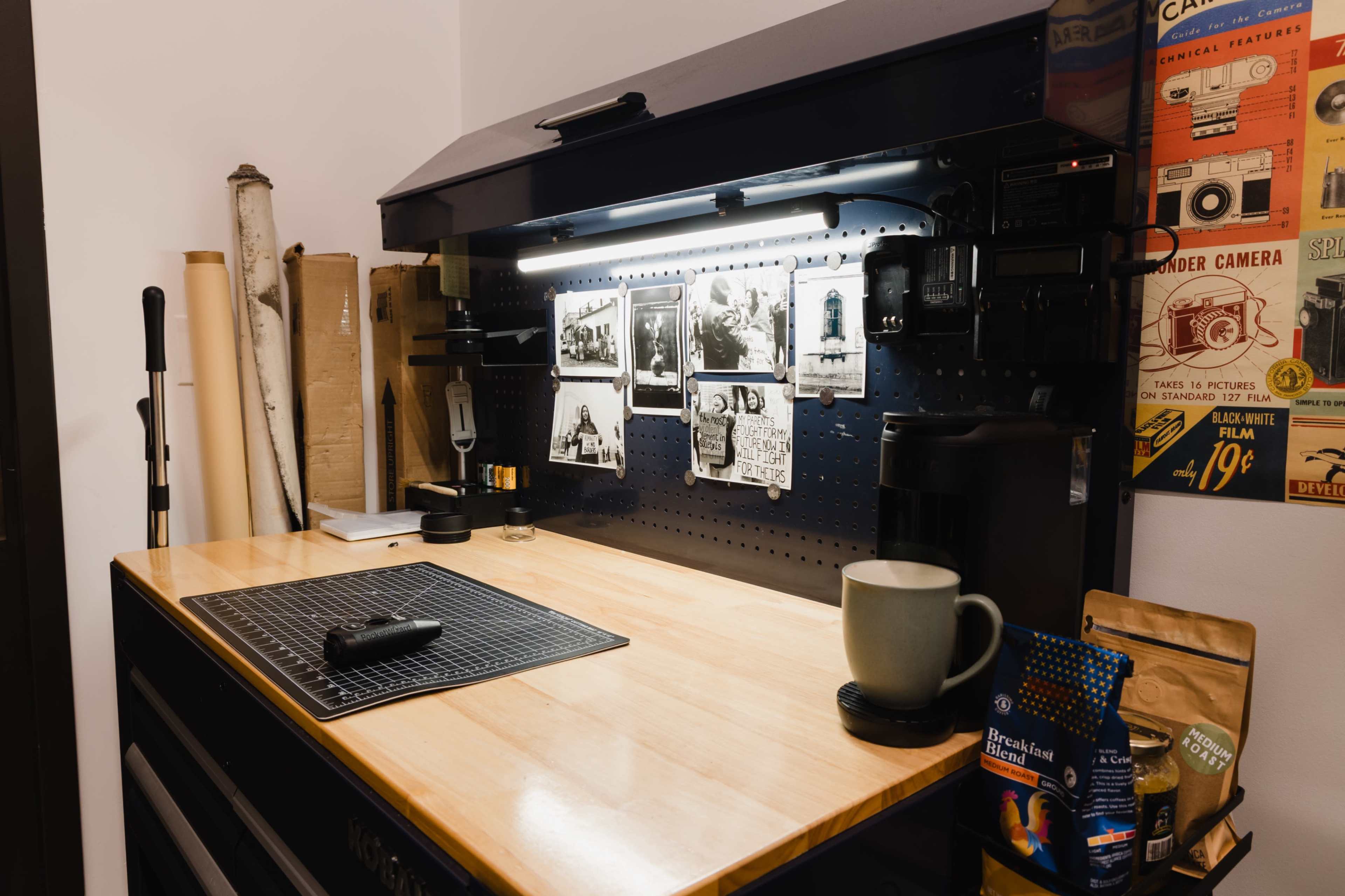 The image shows a workbench with a wooden surface, black pegboard displaying photographs, and various tools, along with a coffee maker and a mug.