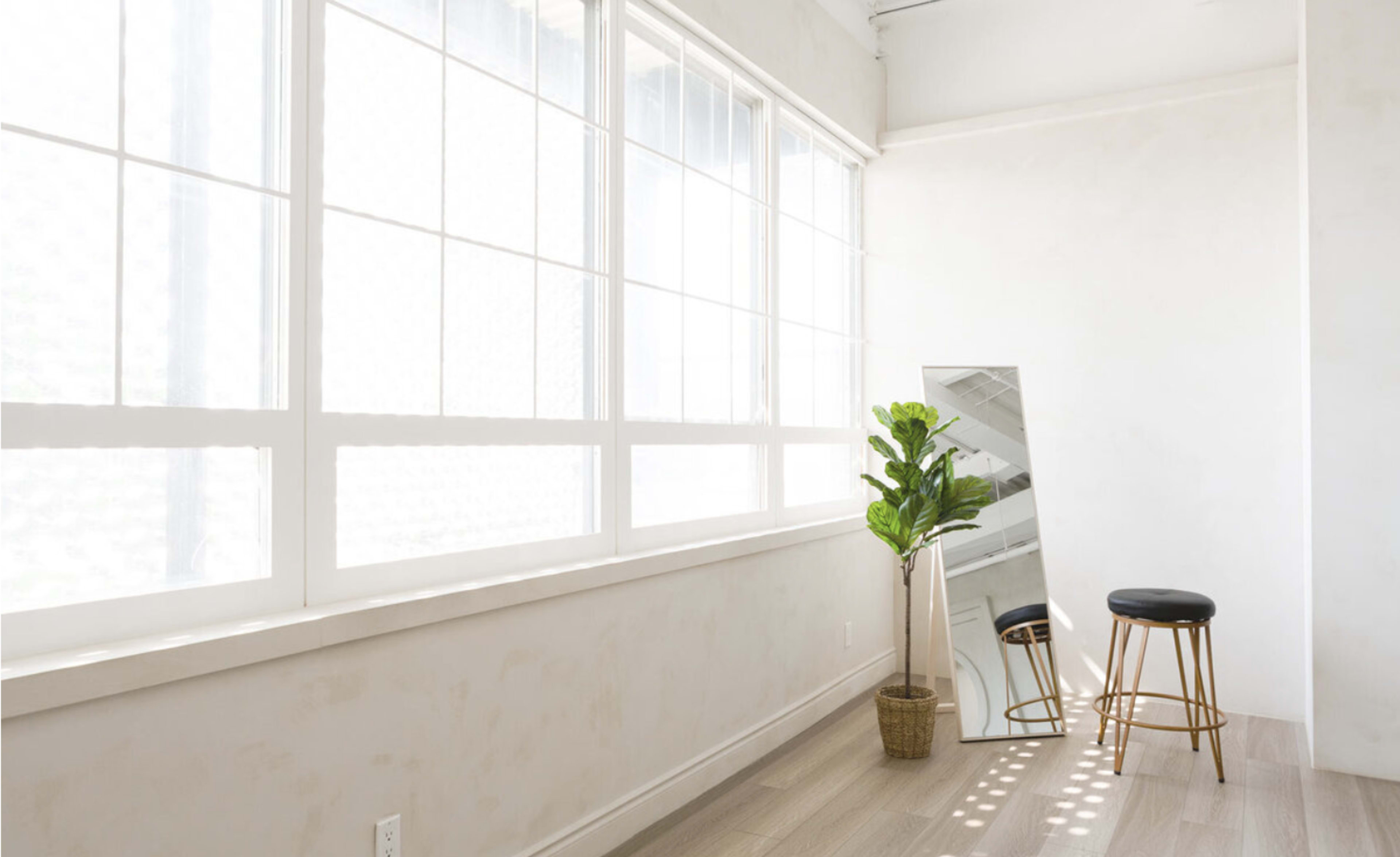 A bright, minimalist room with large windows, a standing mirror, a small potted plant, and a black stool.