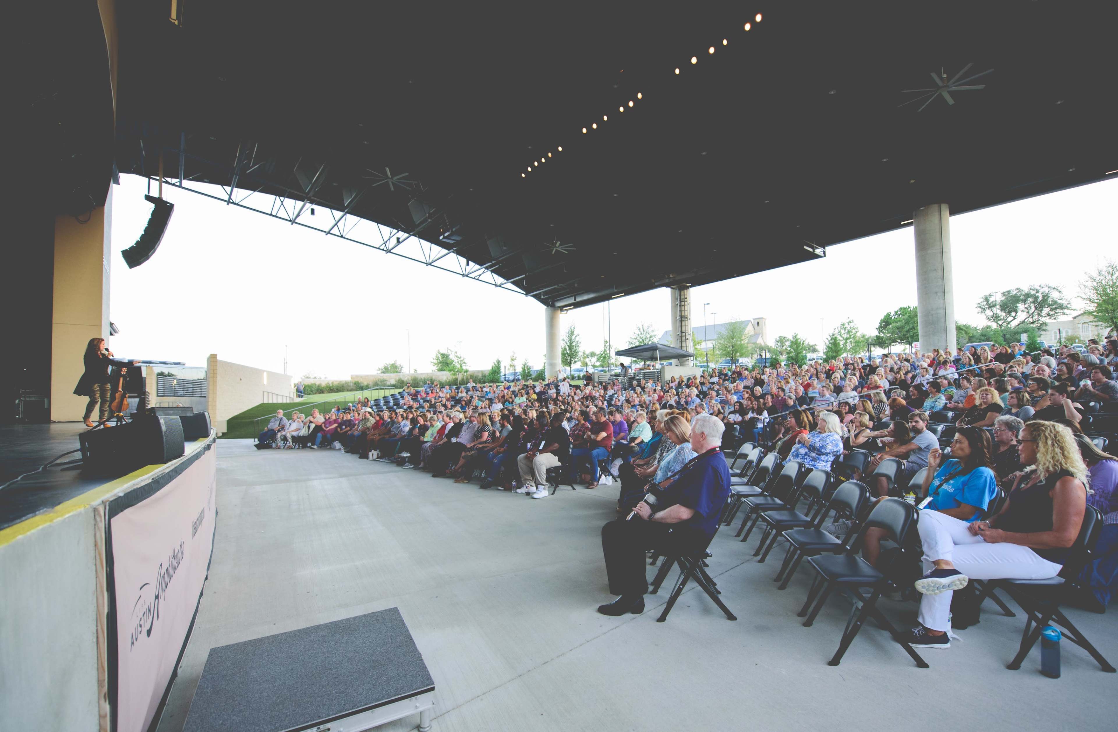 A large audience sits in an amphitheater, watching a speaker on stage under a covered area.