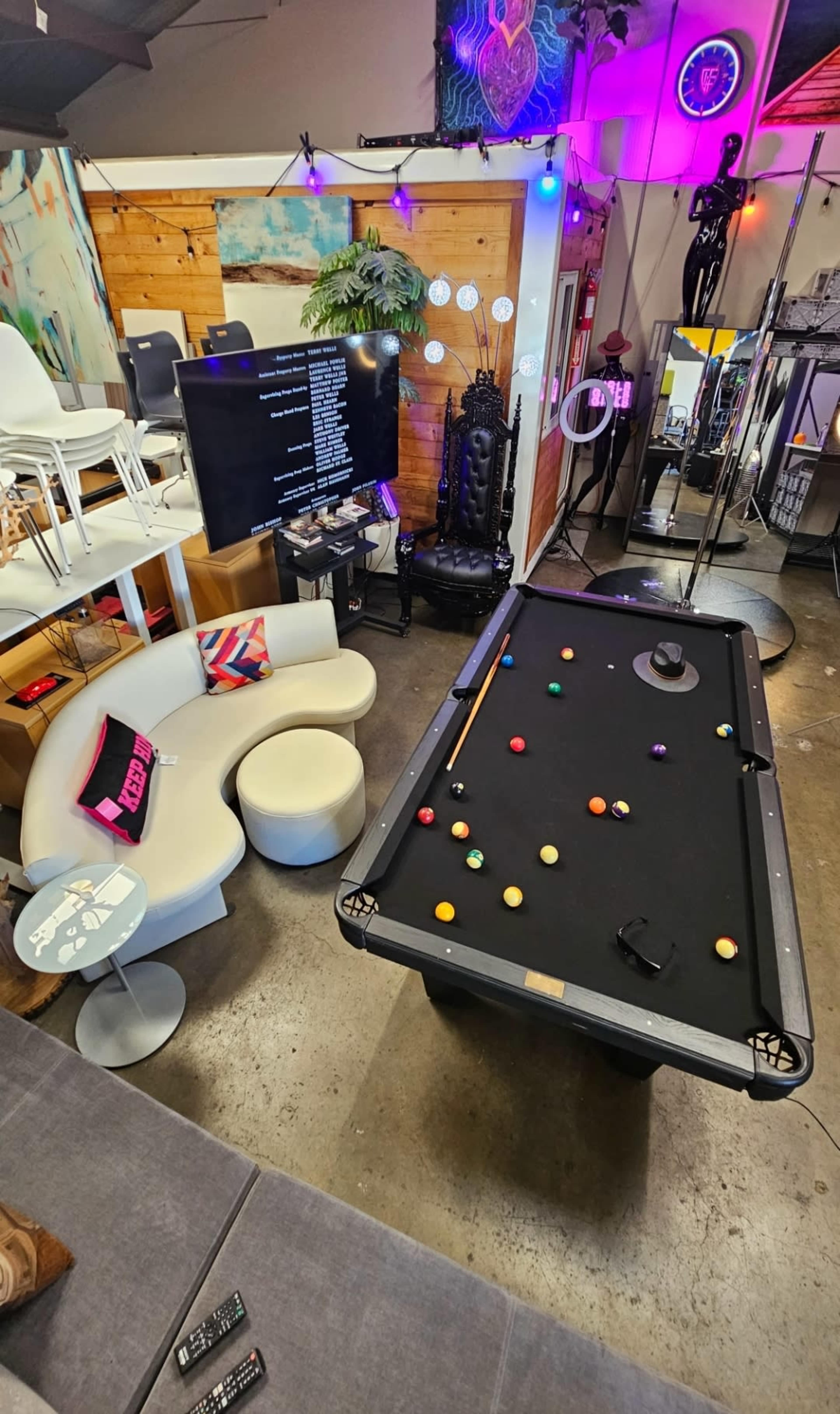 The image shows a recreational space with a pool table in the center, surrounded by seating areas and decor, including a television and various lighting elements.