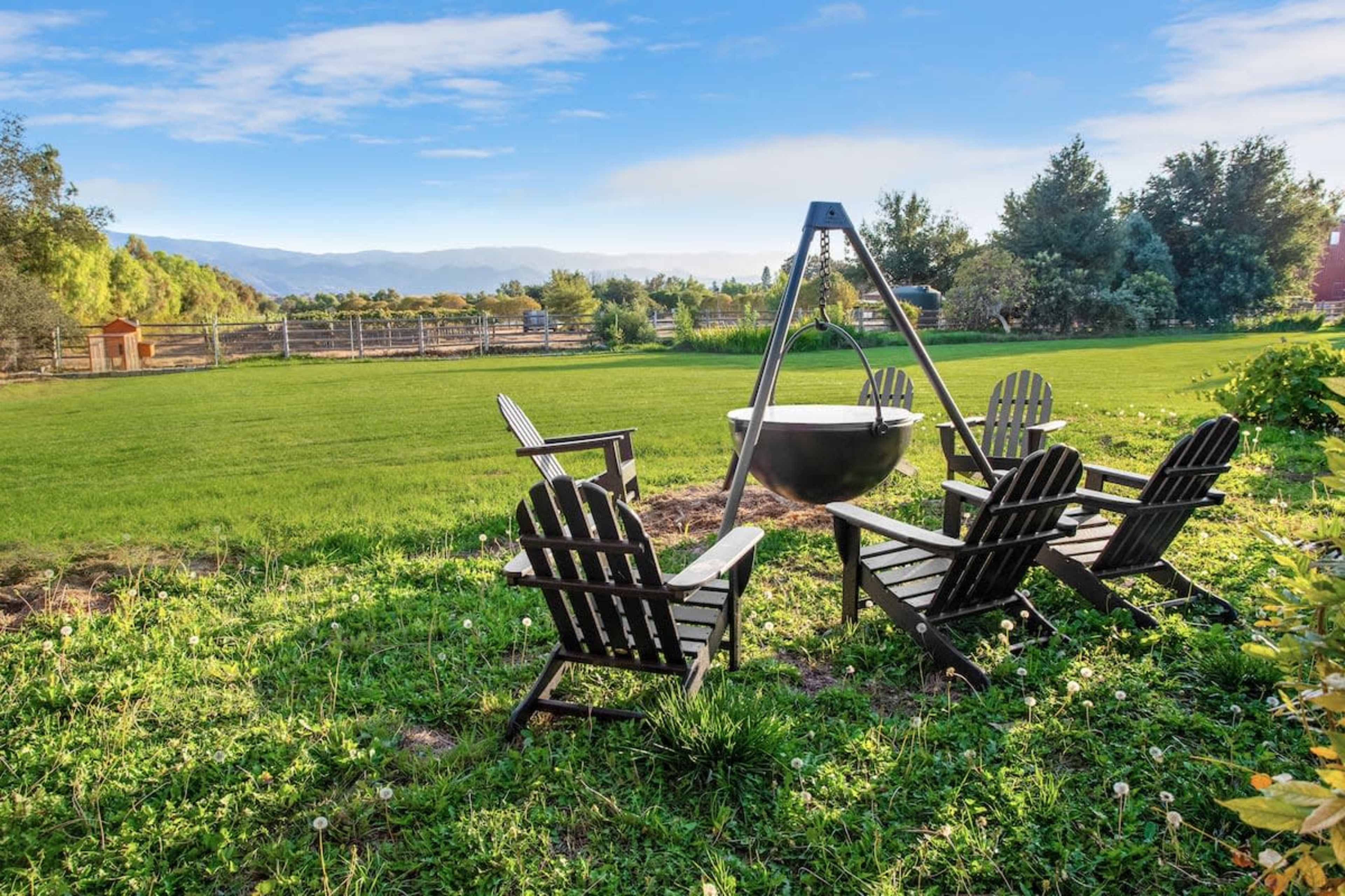 A set of black Adirondack chairs surrounds a round fire pit in a grassy backyard with mountains in the background.