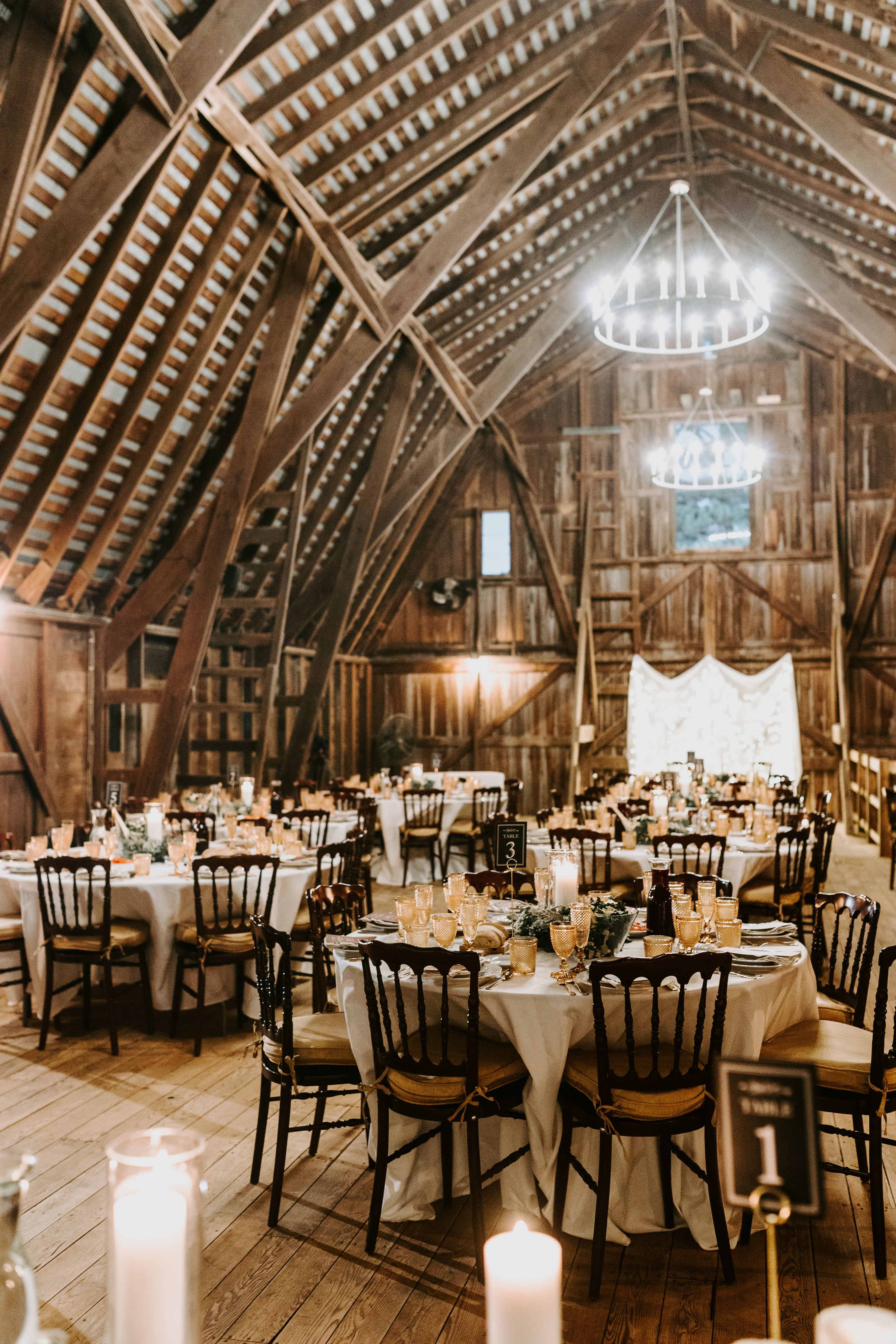 The interior of a rustic barn is set up for a banquet, featuring tables adorned with white linens and centerpieces, surrounded by wooden beams and warm lighting.