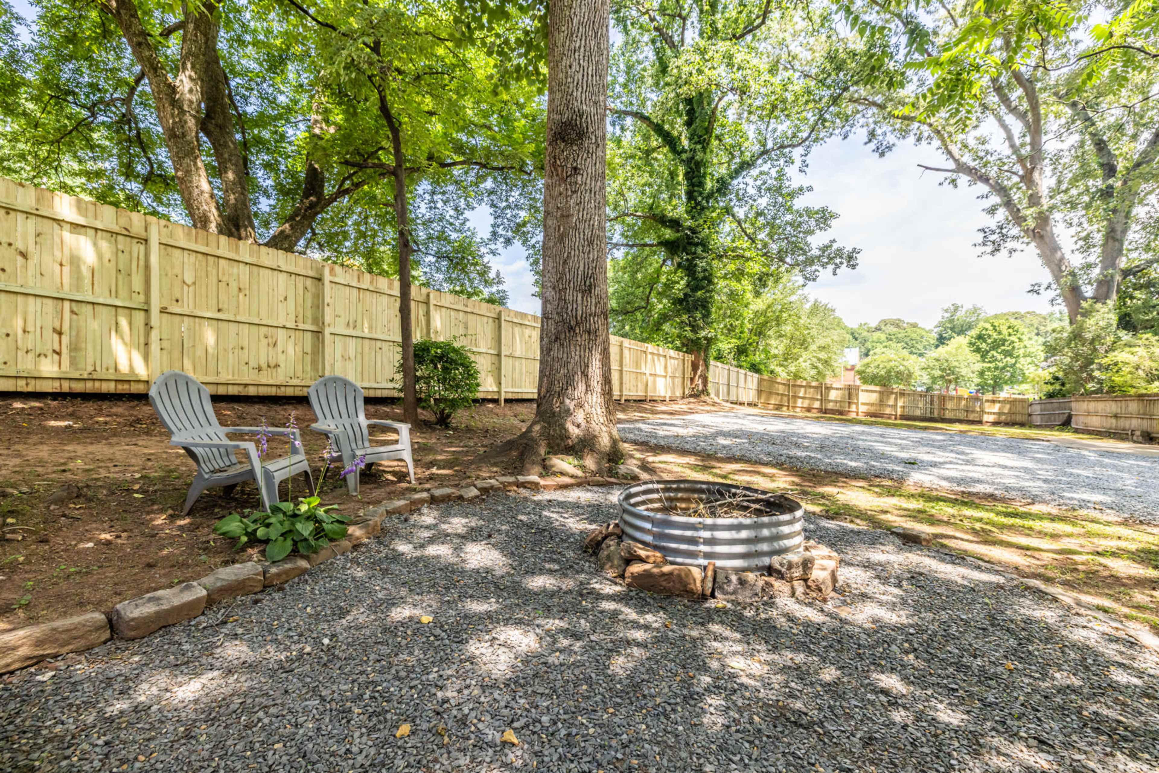 A backyard features a gravel area with a fire pit surrounded by stones and two wooden chairs under the shade of trees, enclosed by a wooden fence.