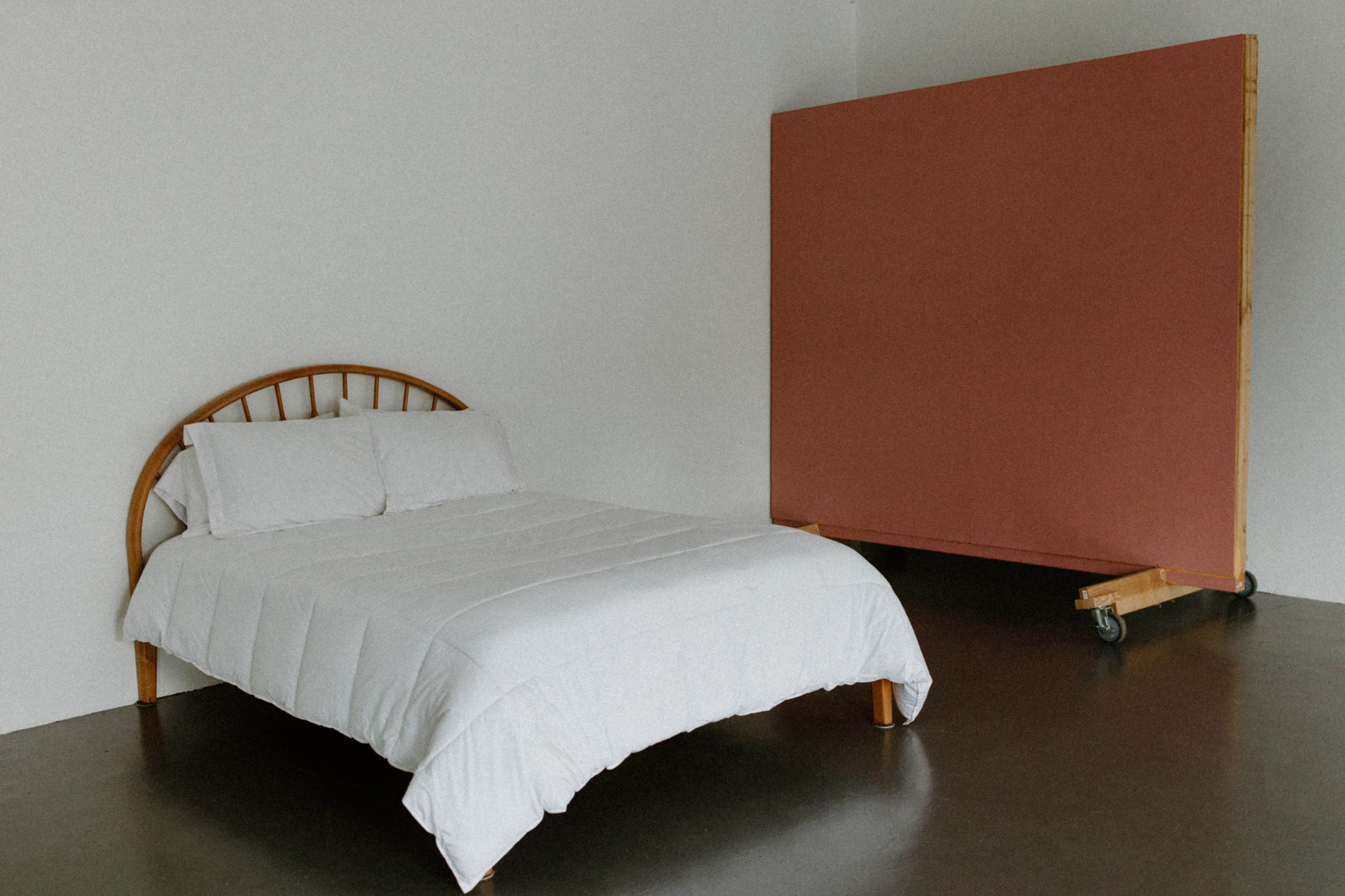A simple bedroom features a wooden bed with a white bedspread and a large pink wall panel on wheels.