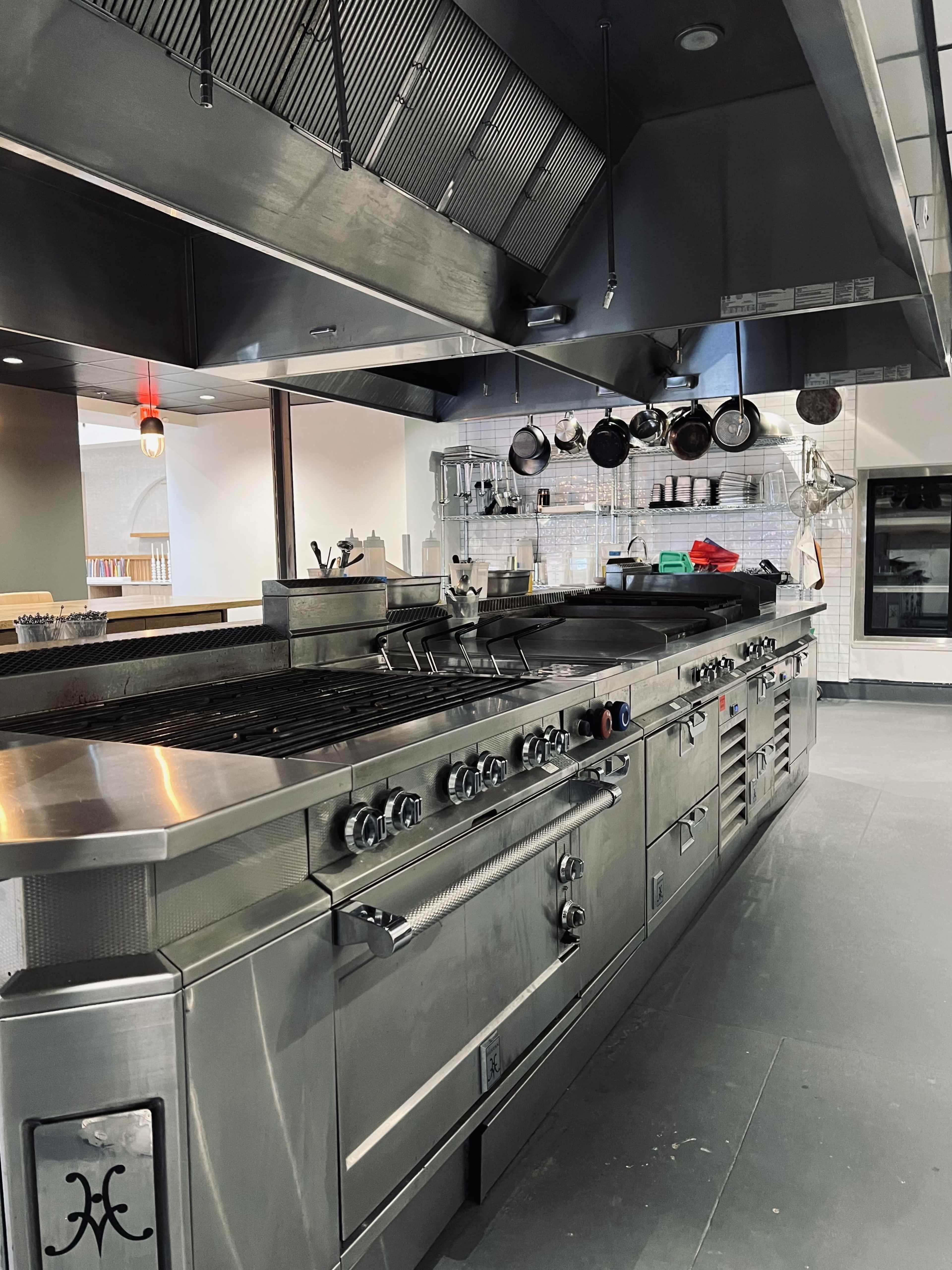 A stainless steel commercial kitchen with a large gas range, overhead ventilation, and various cooking utensils hanging above.