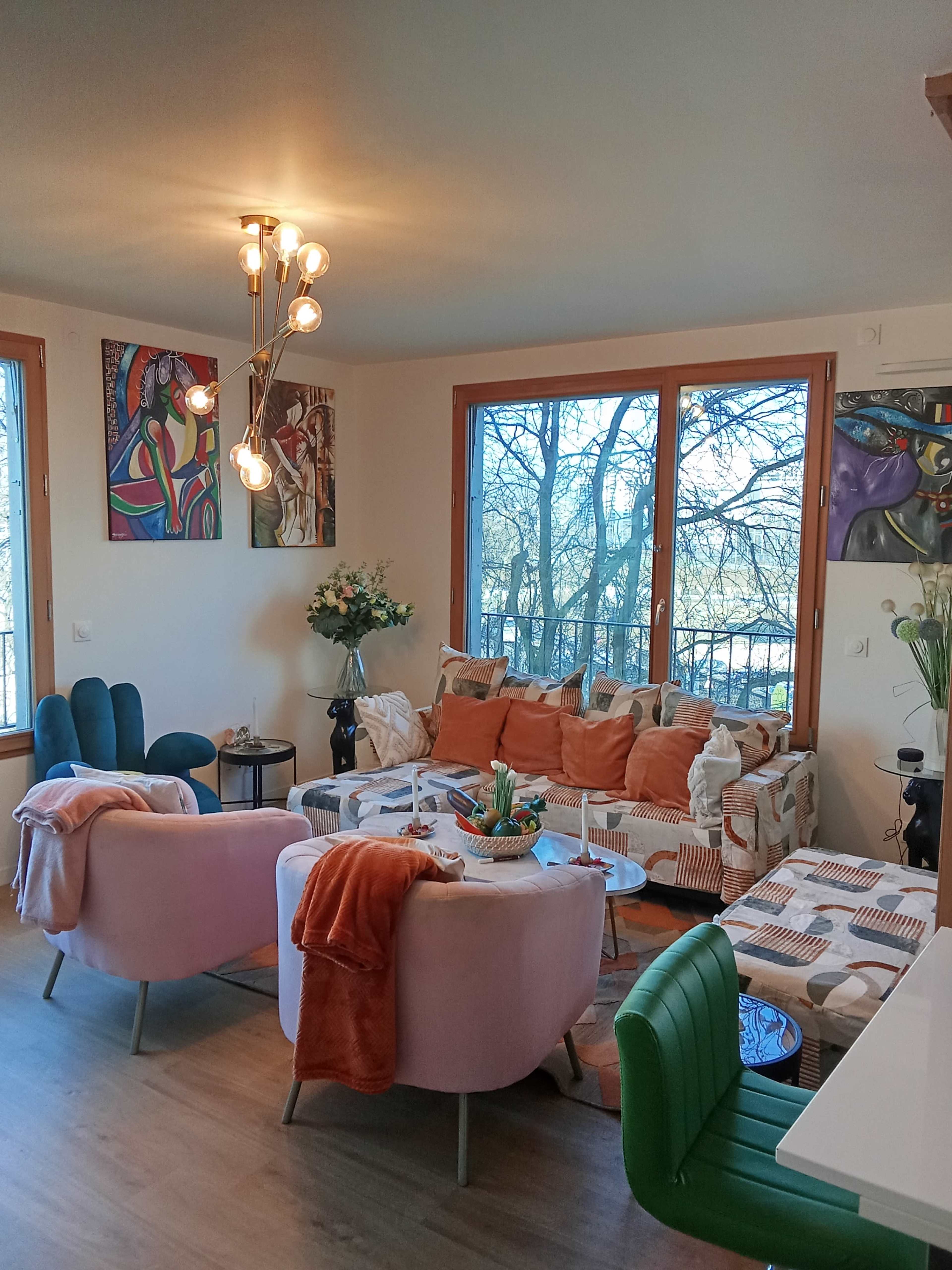 The image shows a cozy living room with a sofa adorned with colorful pillows, a coffee table surrounded by various chairs, and large windows revealing a view of trees outside.