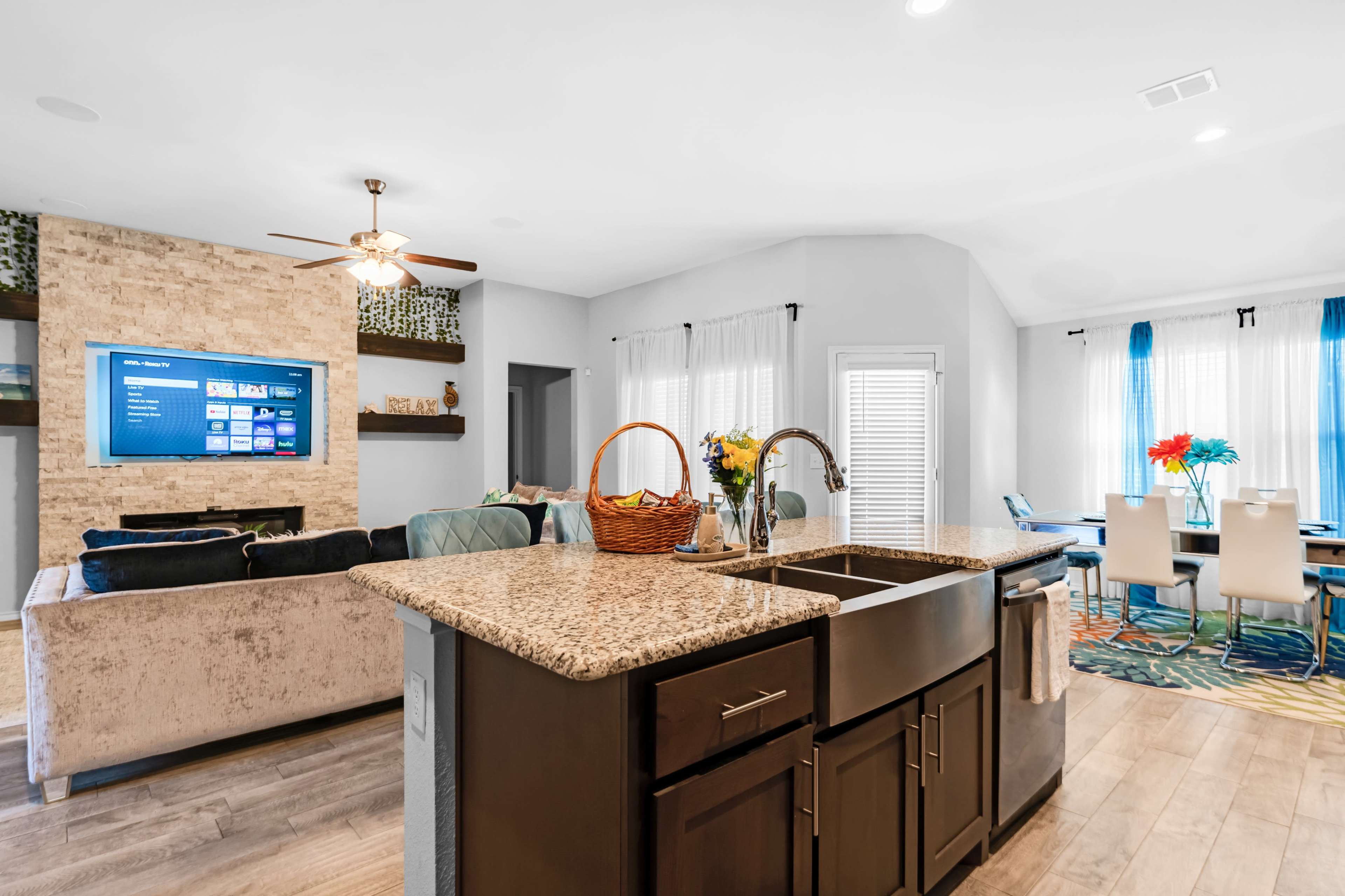A modern kitchen with a granite island, featuring a sink, and an open living area with a wall-mounted TV and a dining space illuminated by natural light.