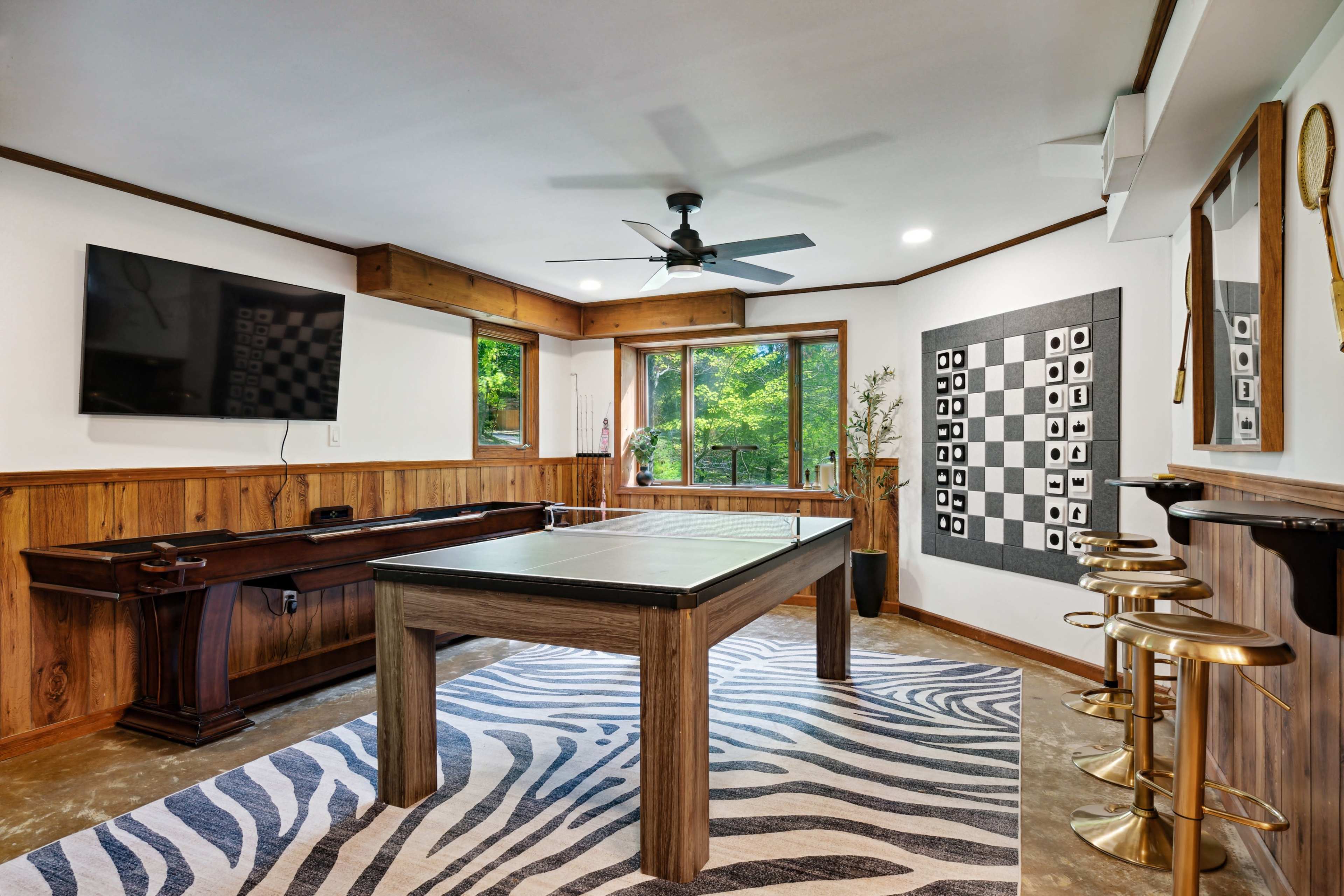 The image shows a modern game room featuring a ping pong table, a television mounted on the wall, and a bar area with gold stools.