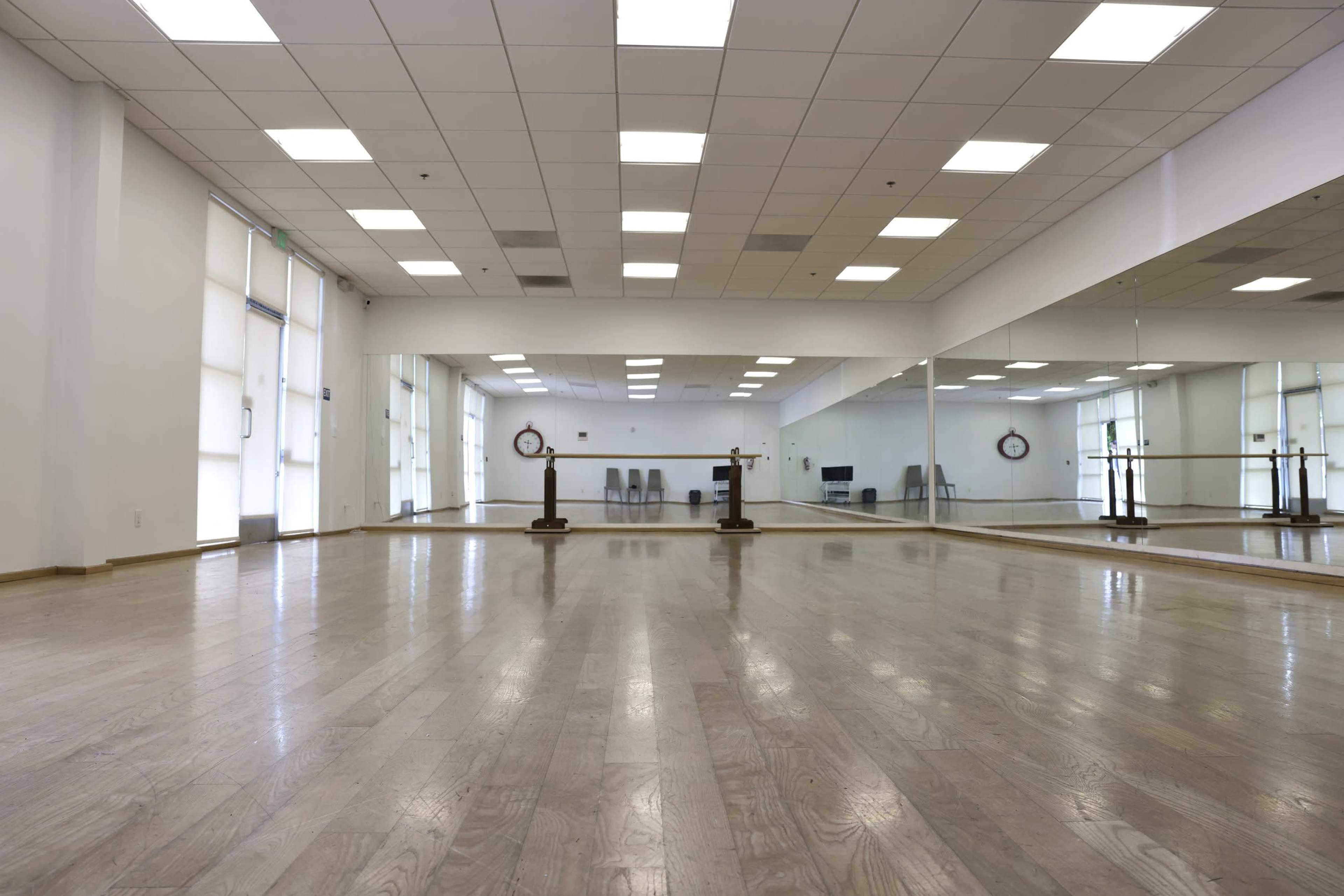 A bright, empty dance studio features wooden flooring, mirrored walls, and a ballet barre.