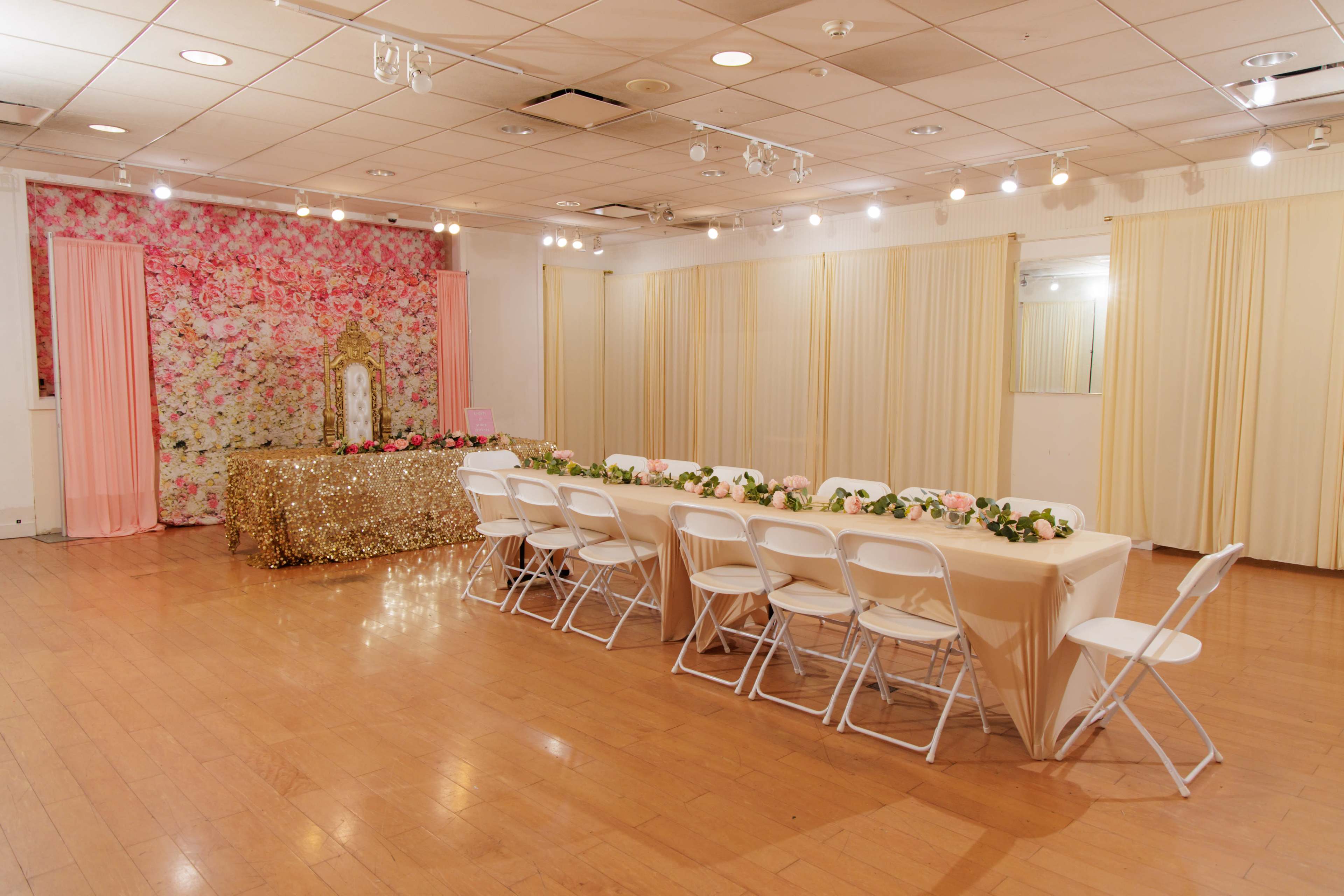 The image shows a decorated event space featuring a long table with white chairs, a floral backdrop, and a gold sequined tablecloth.