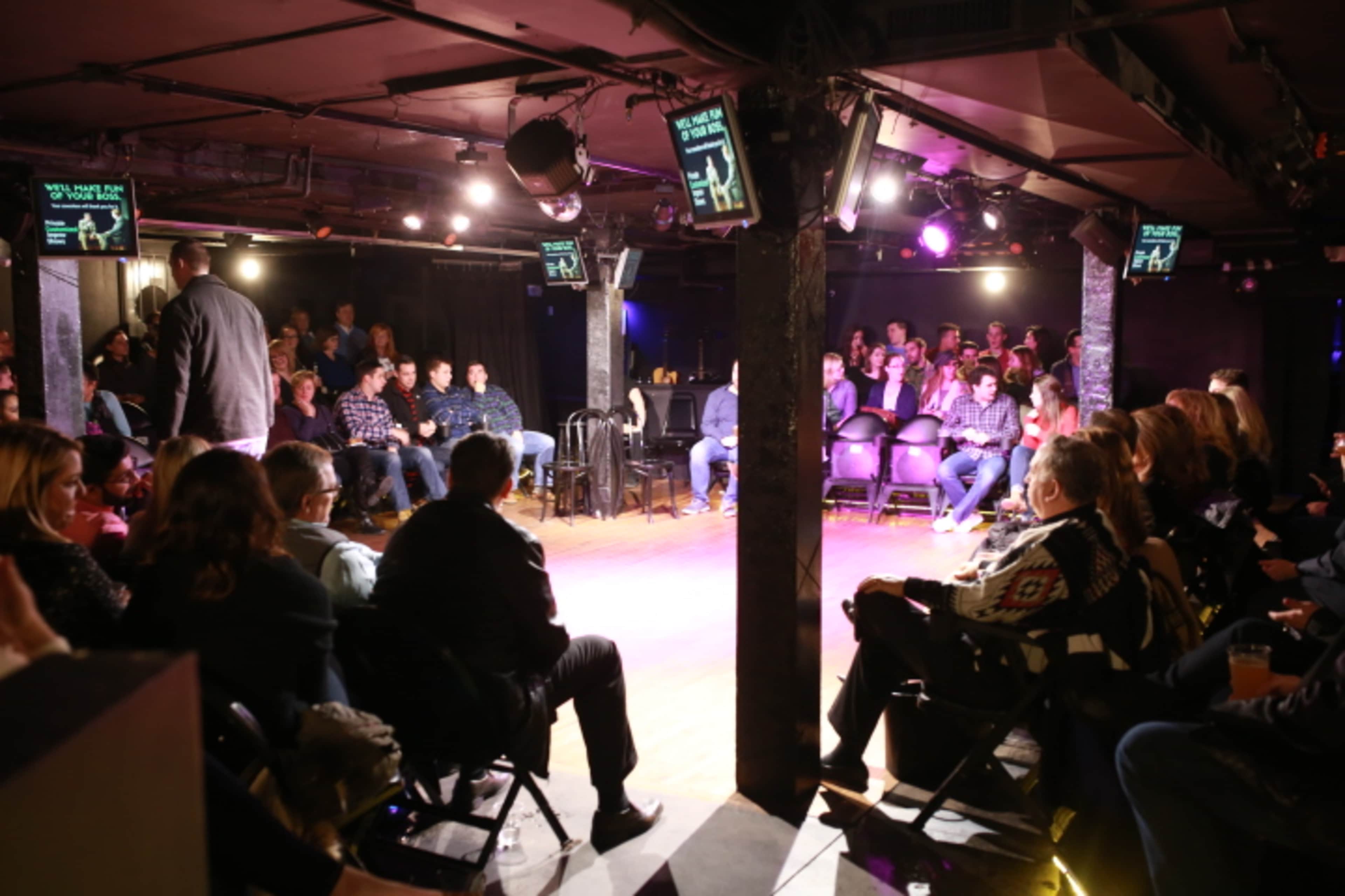 A comedy club audience is seated in rows facing a small stage with multiple screens displaying information.