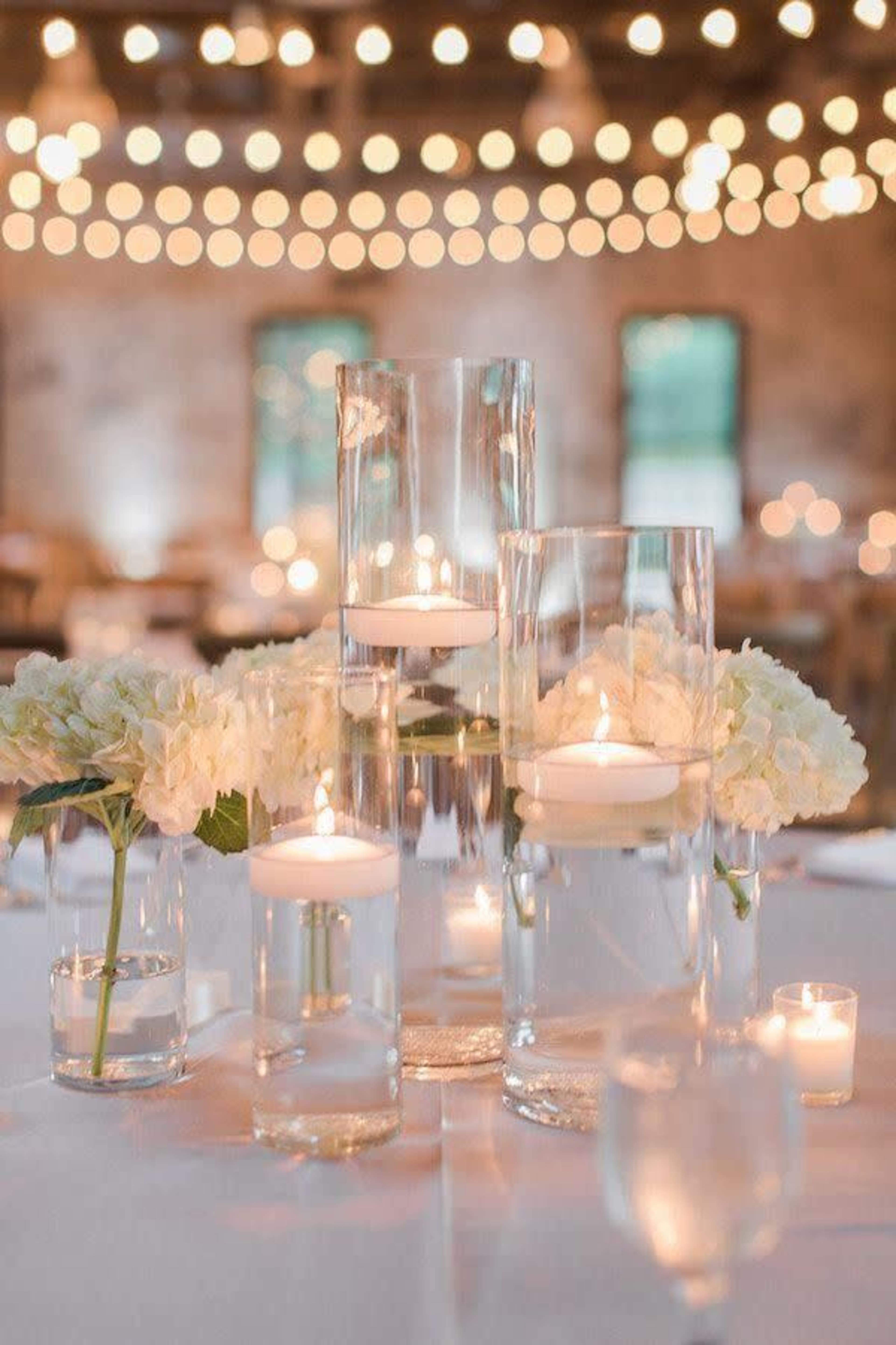 A table setting adorned with glass vases of varying heights, each containing floating candles and surrounded by white hydrangeas, all illuminated by soft, warm string lights in the background.