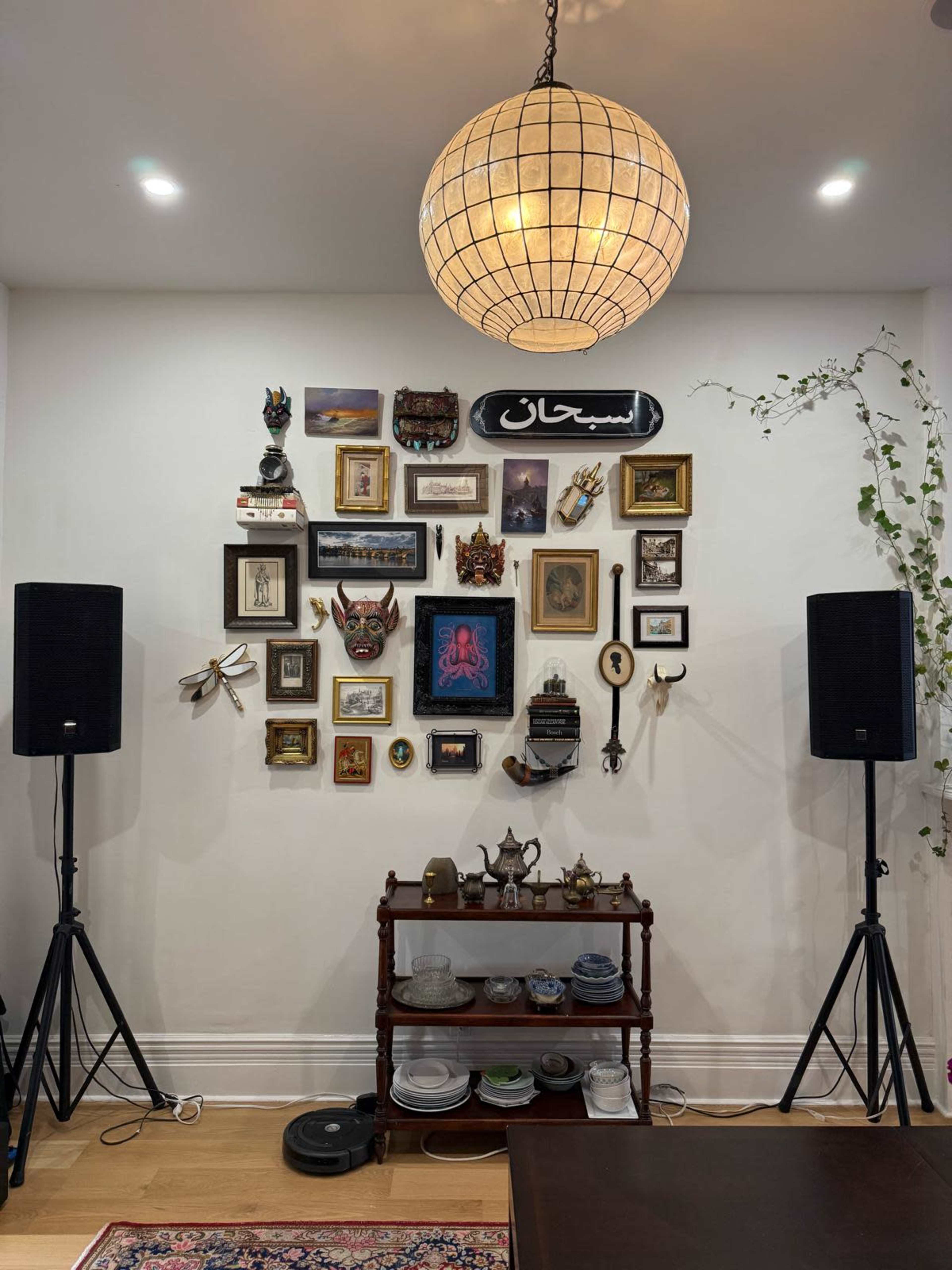 The image shows a decorated wall featuring various framed artworks and artifacts, flanked by two large speakers, with a wooden shelf holding plates and other decorative items beneath.