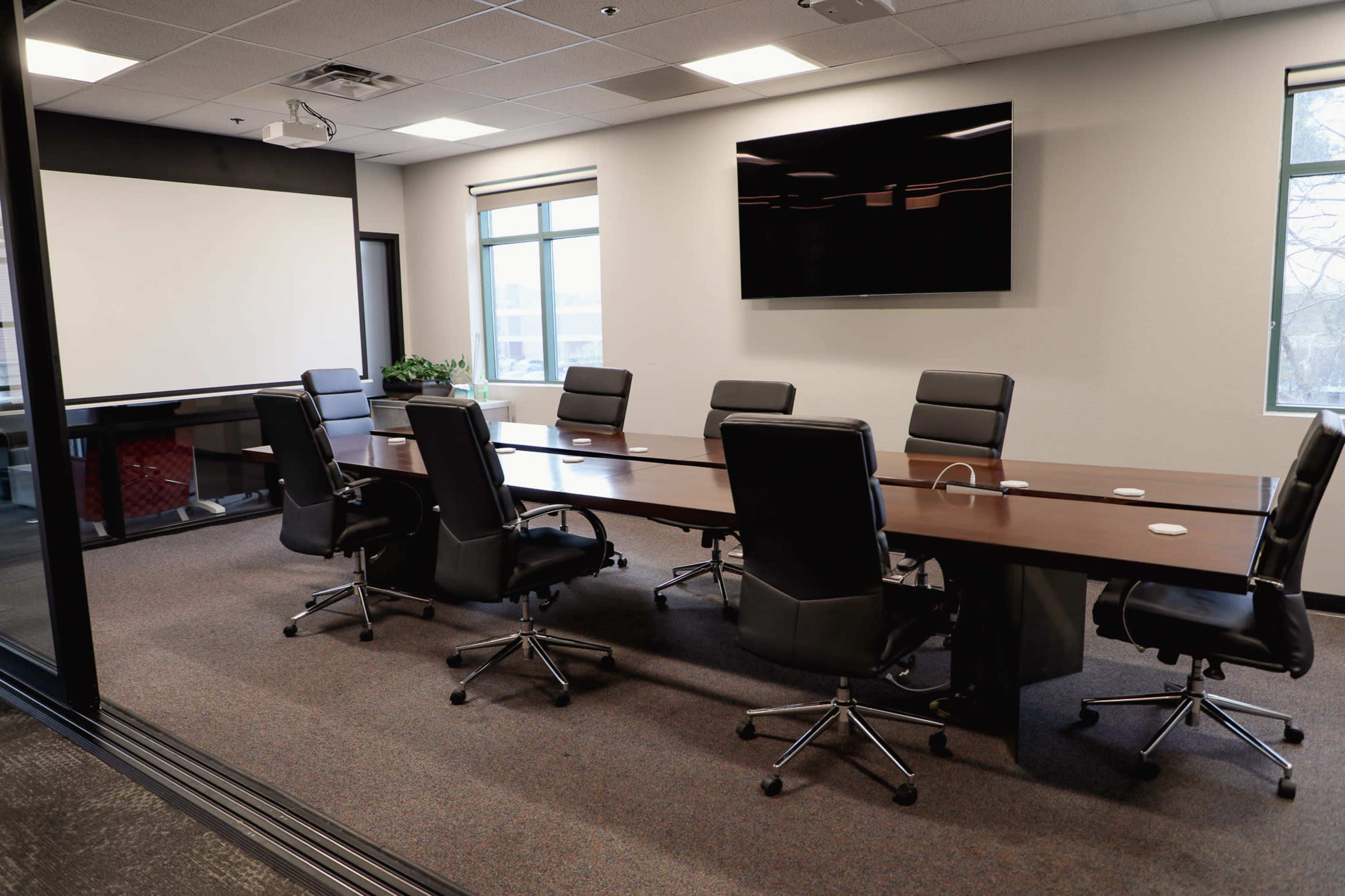 A modern conference room features a long wooden table surrounded by black rolling chairs, with a large screen mounted on the wall and windows allowing natural light.