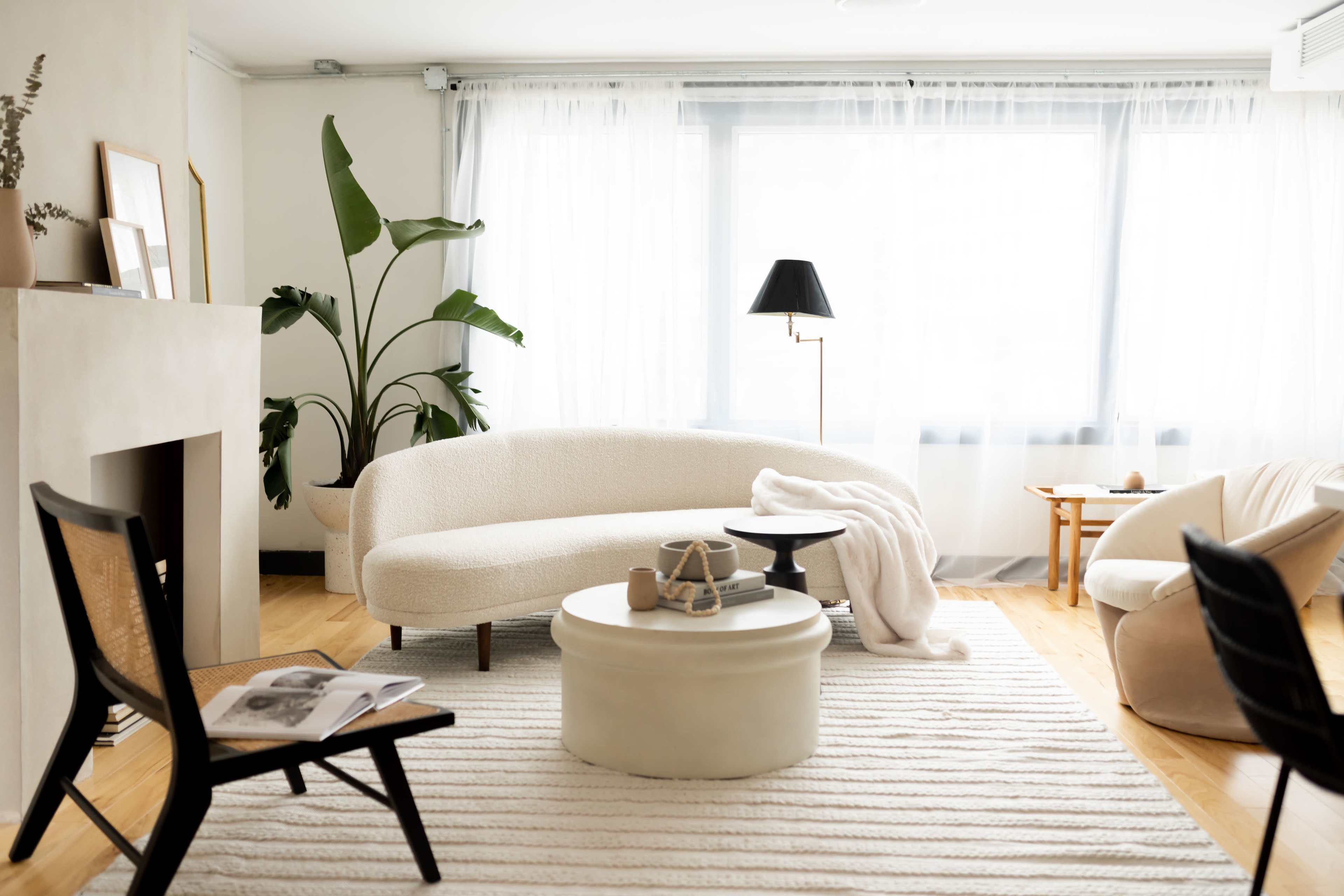 A modern living room features a curved white sofa, a round coffee table, and various seating options, with large windows allowing natural light.