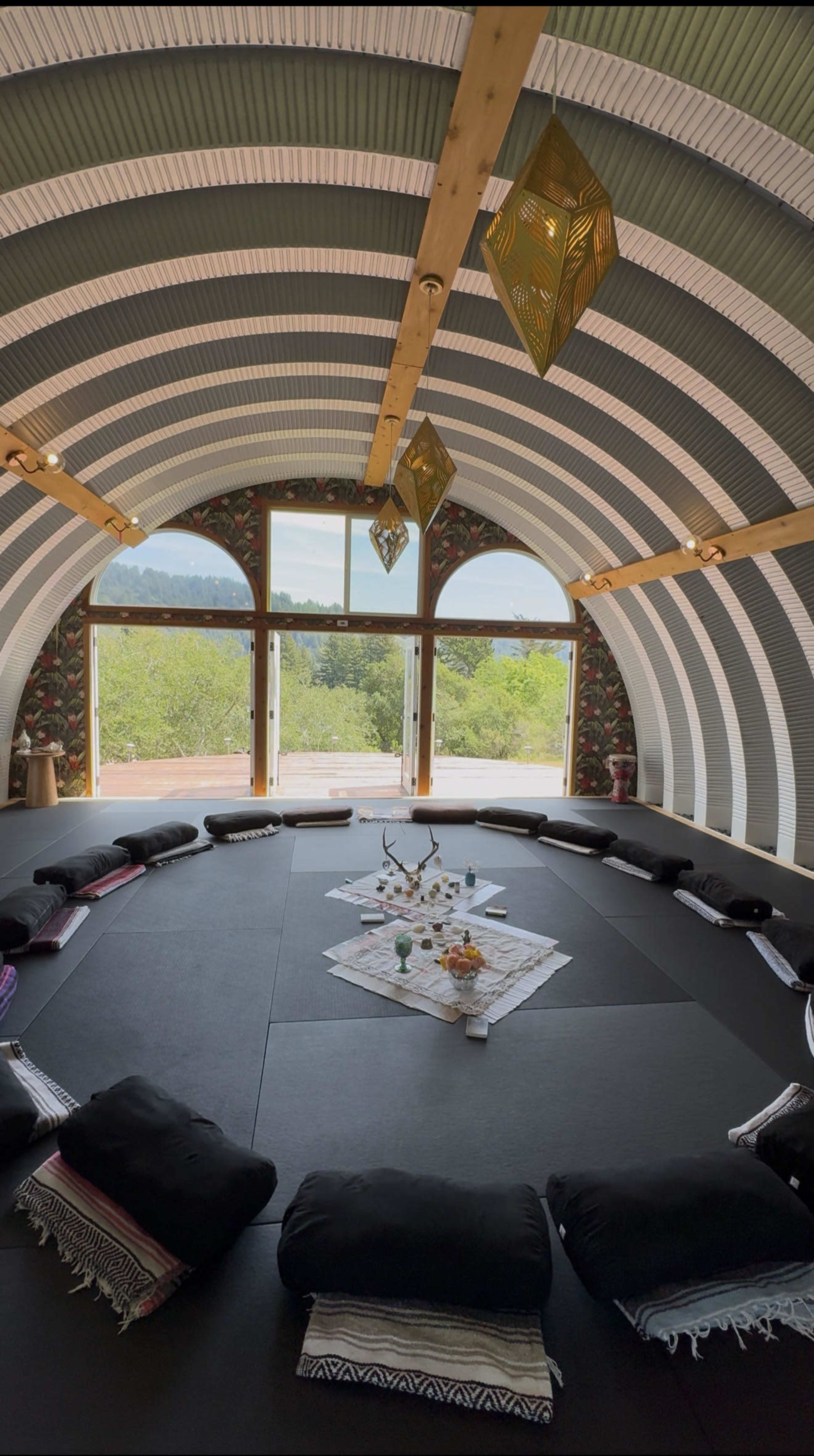 The image shows a circular room with a black mat floor, surrounded by pillows arranged around a central decorative setup on a table, and large windows providing a view of greenery outside.
