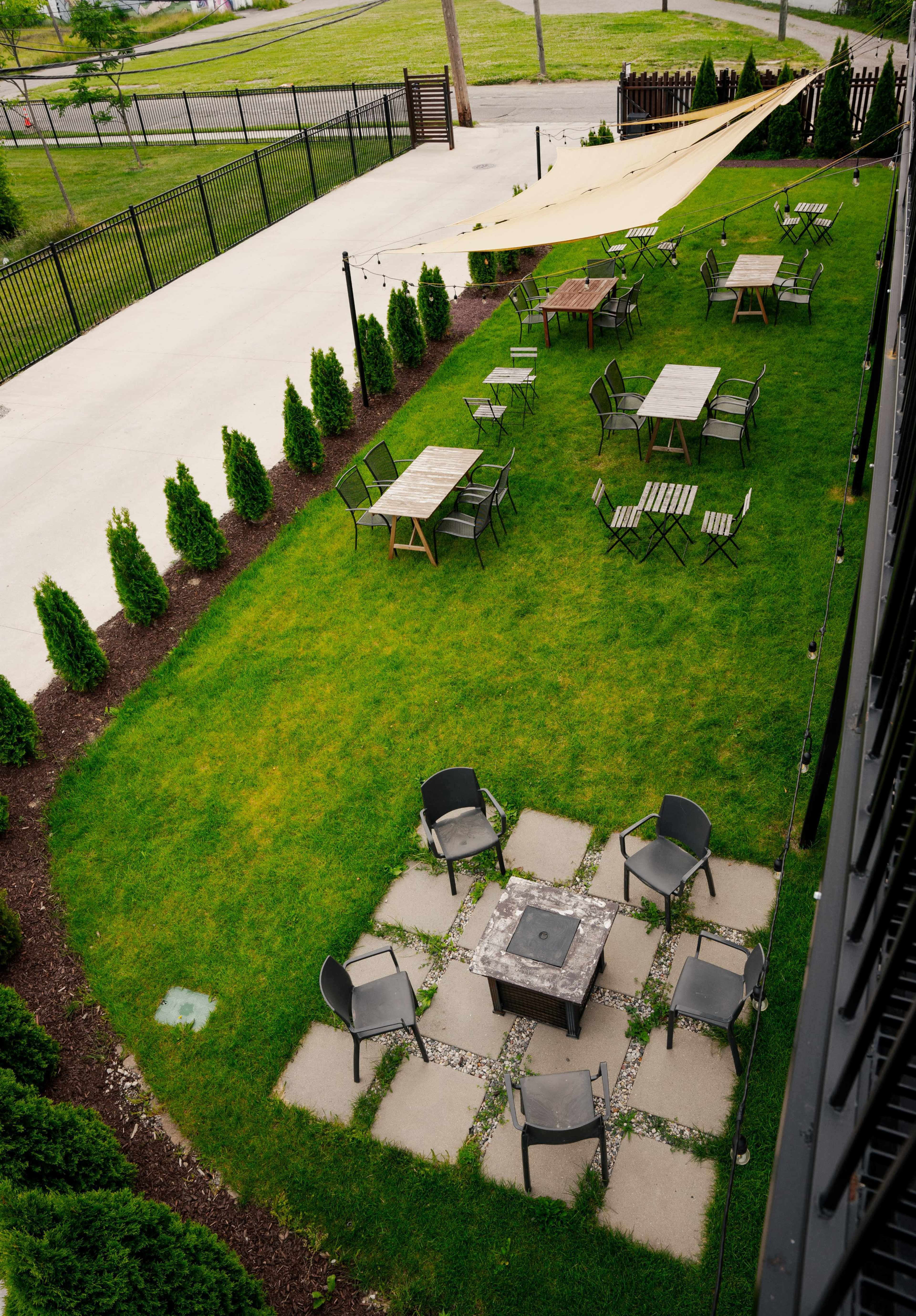 A landscaped outdoor area with several tables and chairs arranged on a lawn, alongside neatly trimmed hedges and a fire pit surrounded by stone pavers.