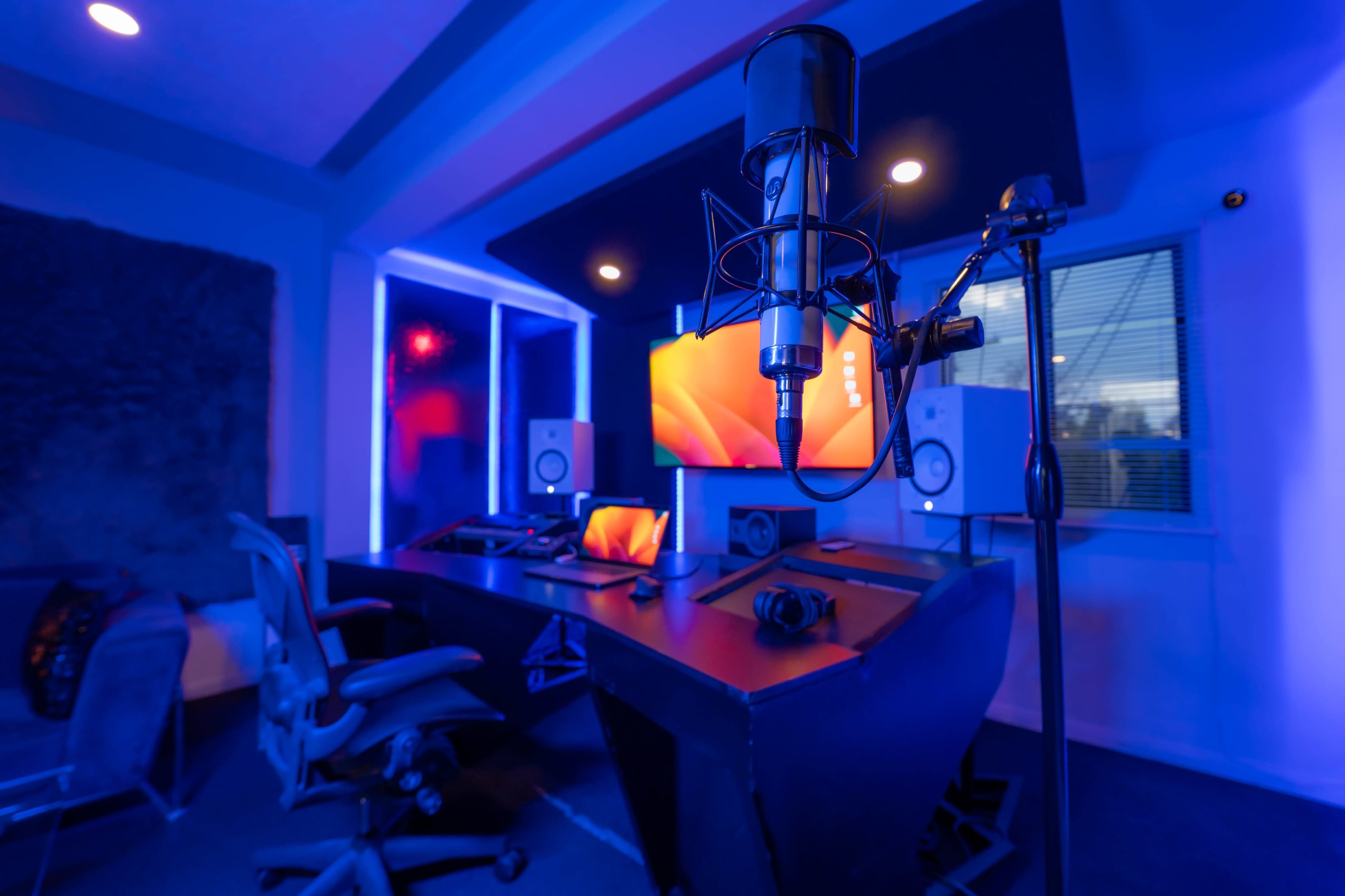 A modern music studio features a microphone stationed in front of a computer setup, surrounded by blue lighting and various audio equipment.