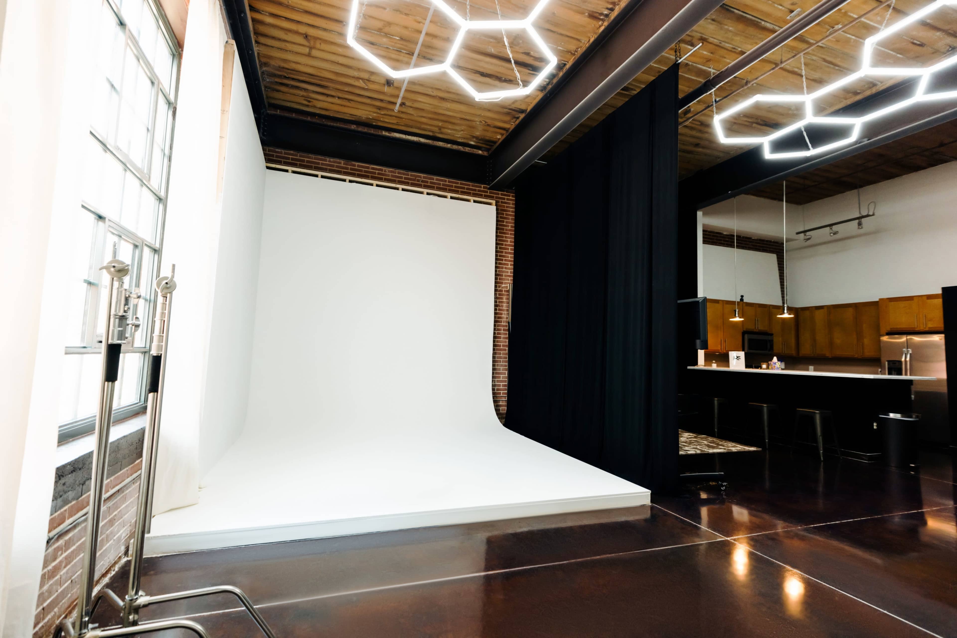 The image shows a spacious studio interior with a white photography backdrop, a black curtain, and a kitchen area with wooden cabinets and modern lighting fixtures.