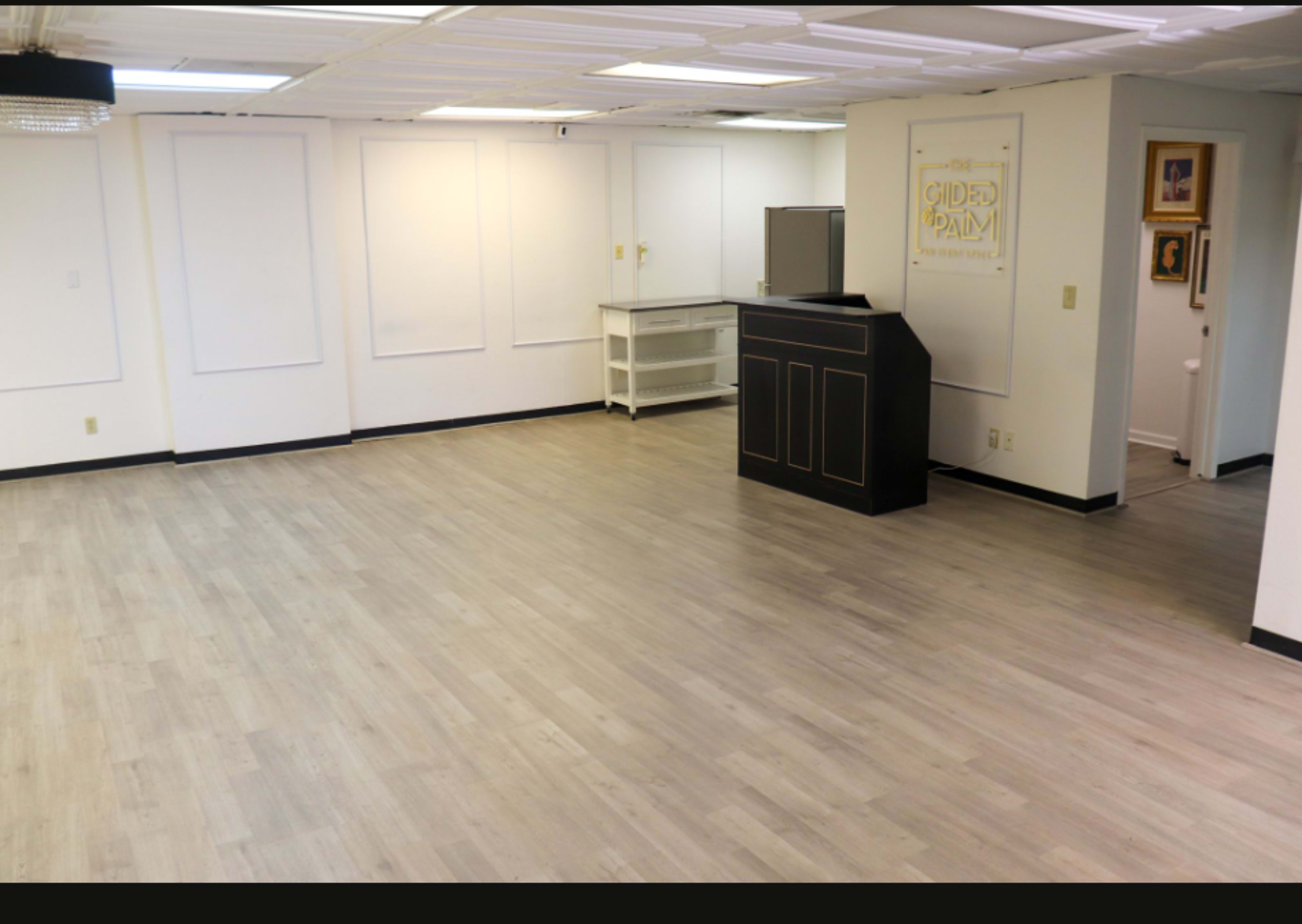 The image shows an empty room with light-colored wooden flooring, white walls, and a reception area consisting of a black counter and a small shelf.