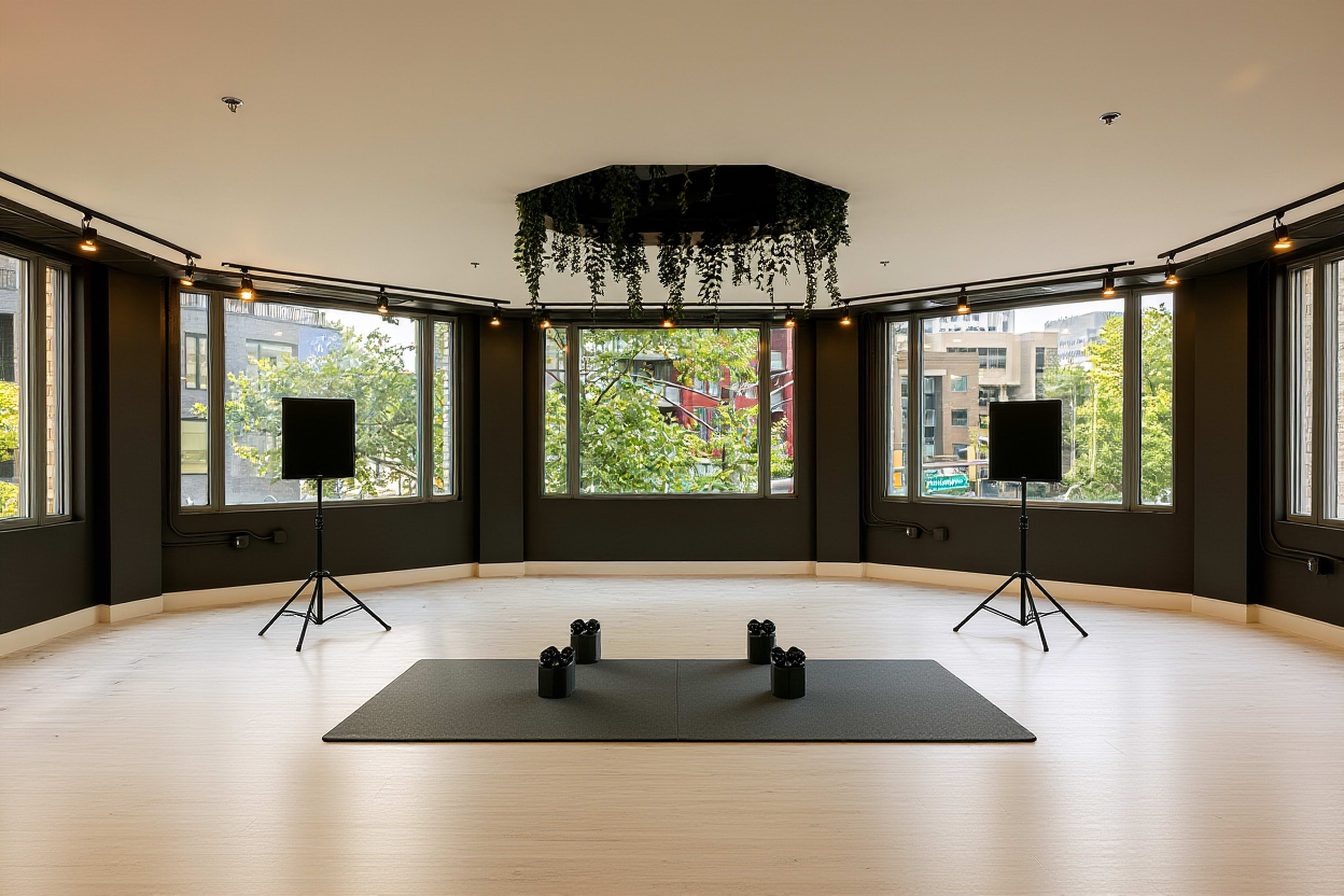 The image shows a spacious room with large windows, featuring a central exercise mat and two speaker stands positioned on either side.