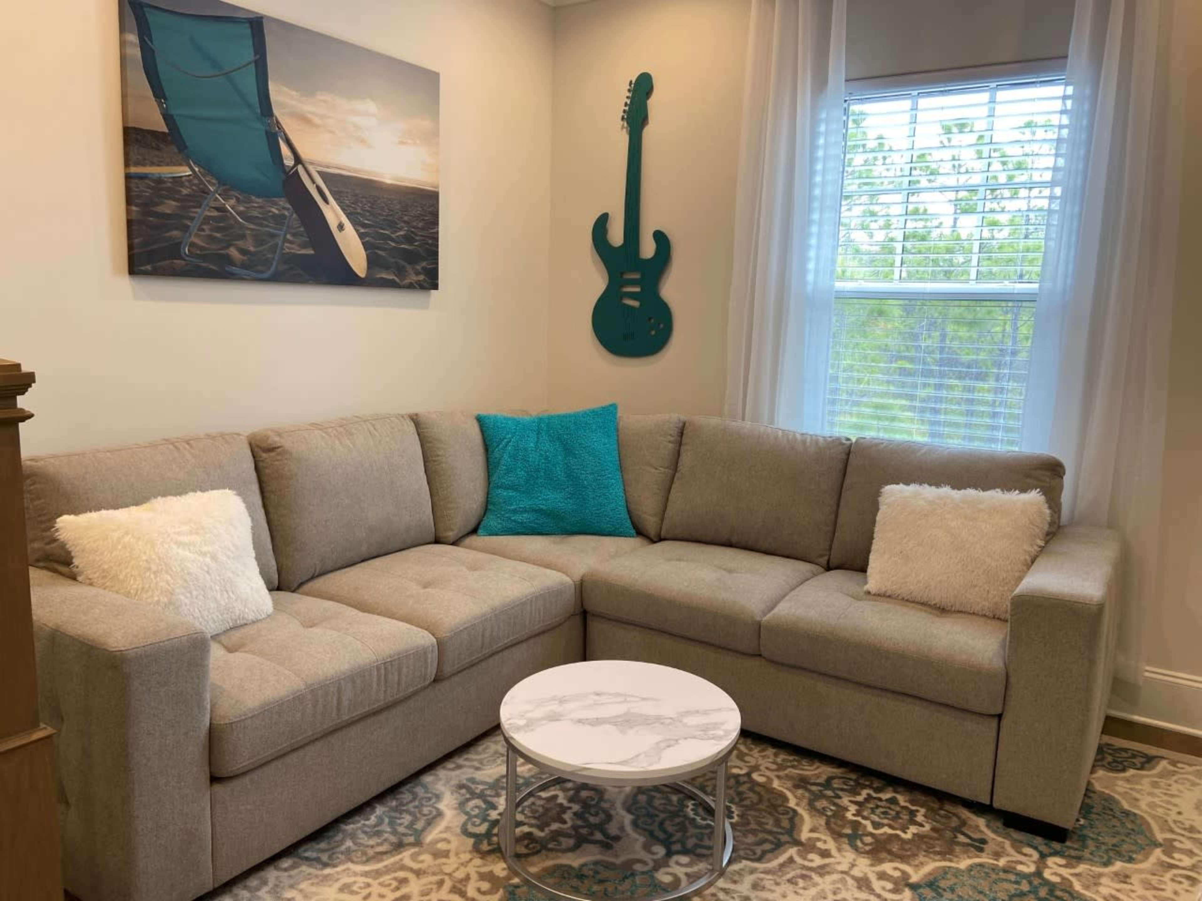 A light-colored sectional sofa with decorative pillows is positioned in a room featuring a teal guitar on the wall and a round marble coffee table.