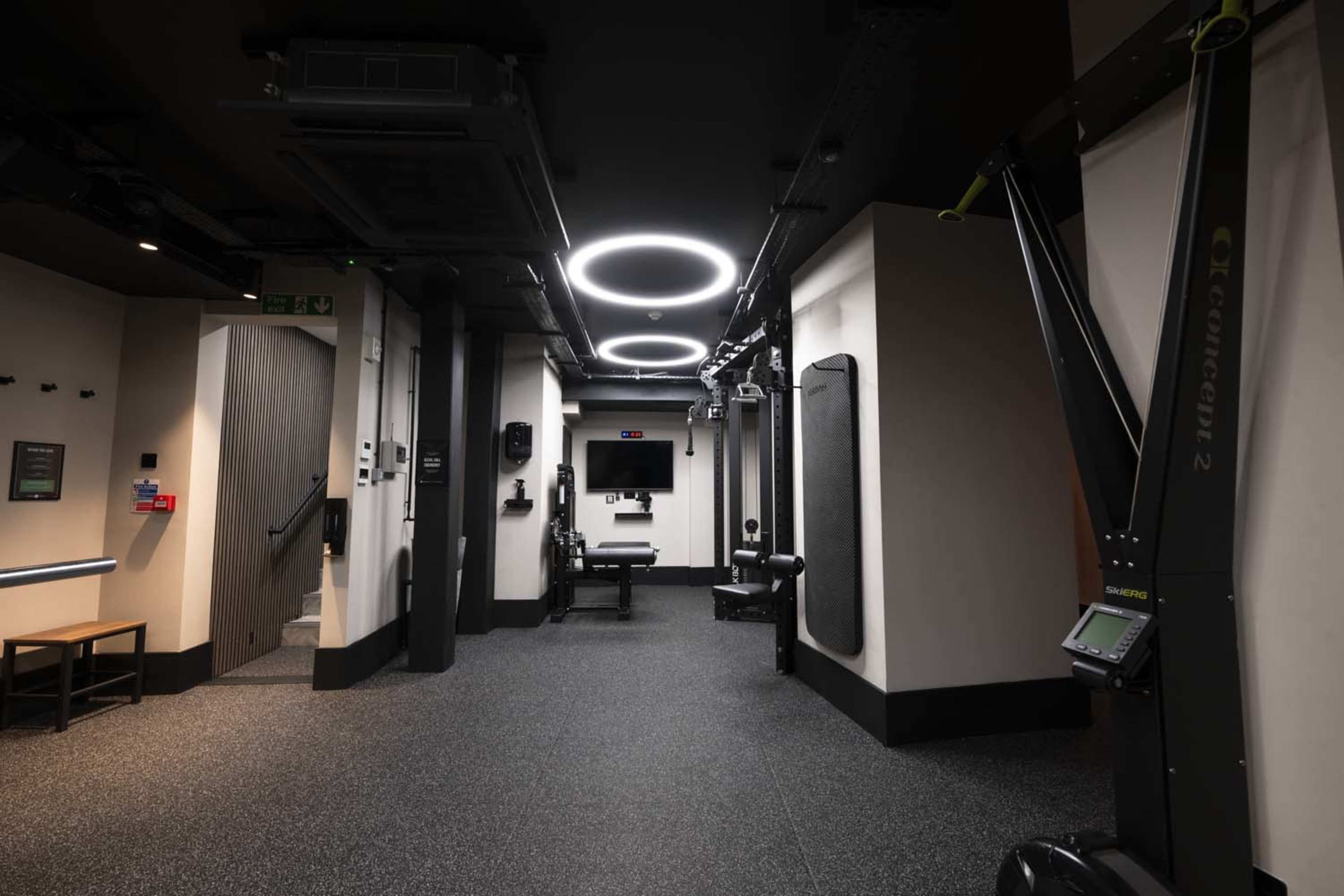 The image shows a modern gym space with rubber flooring, various exercise equipment, and circular LED lighting.