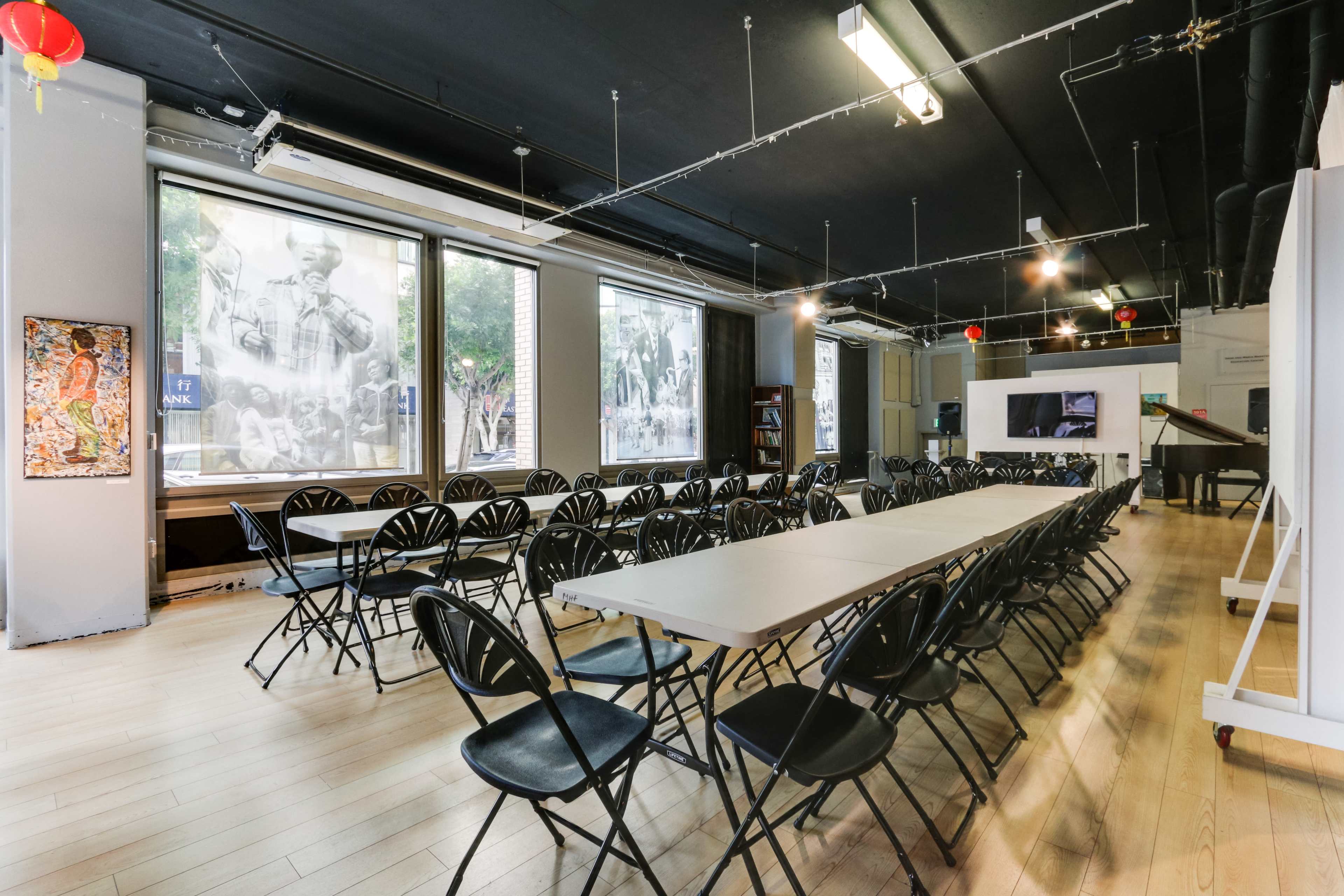A spacious room is set up with rows of folding chairs facing a long table, surrounded by large windows showcasing black-and-white photographs.