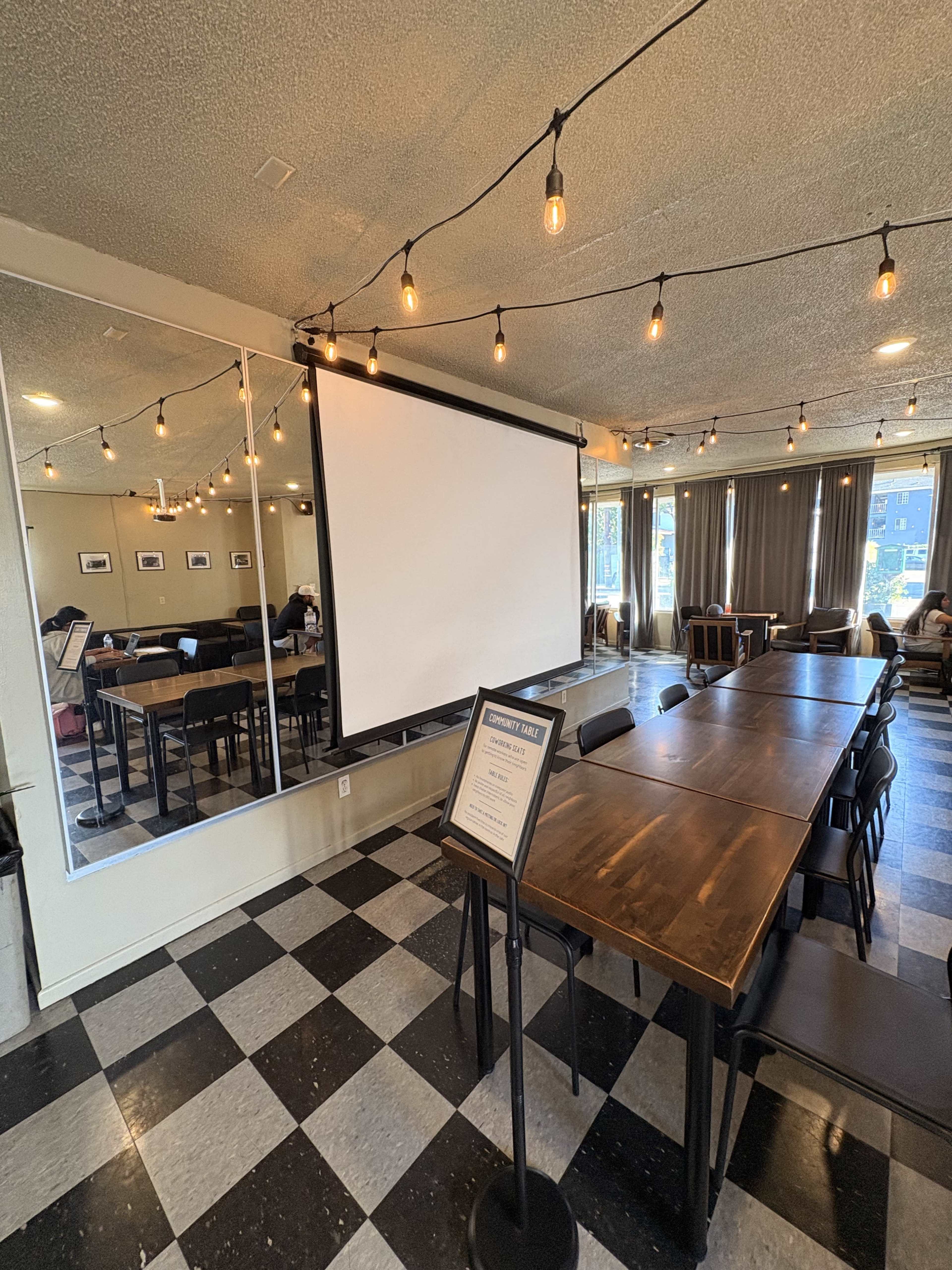 The image shows a conference room with a long wooden table, a projector screen, decorative string lights, and a mirrored wall.