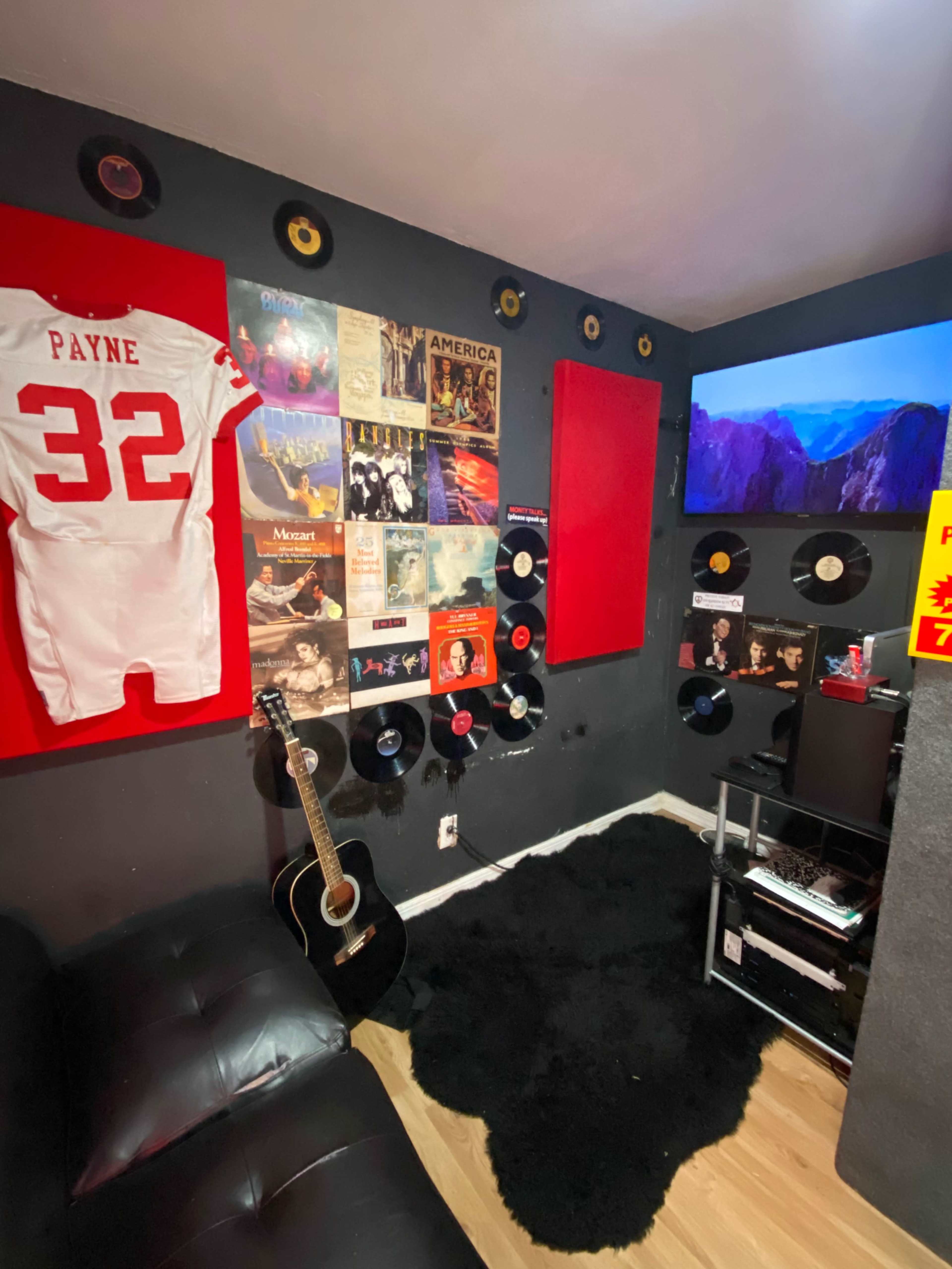 A music-themed room with a black sofa, records mounted on the walls, a guitar resting on the floor, and a television displaying a scenic image.