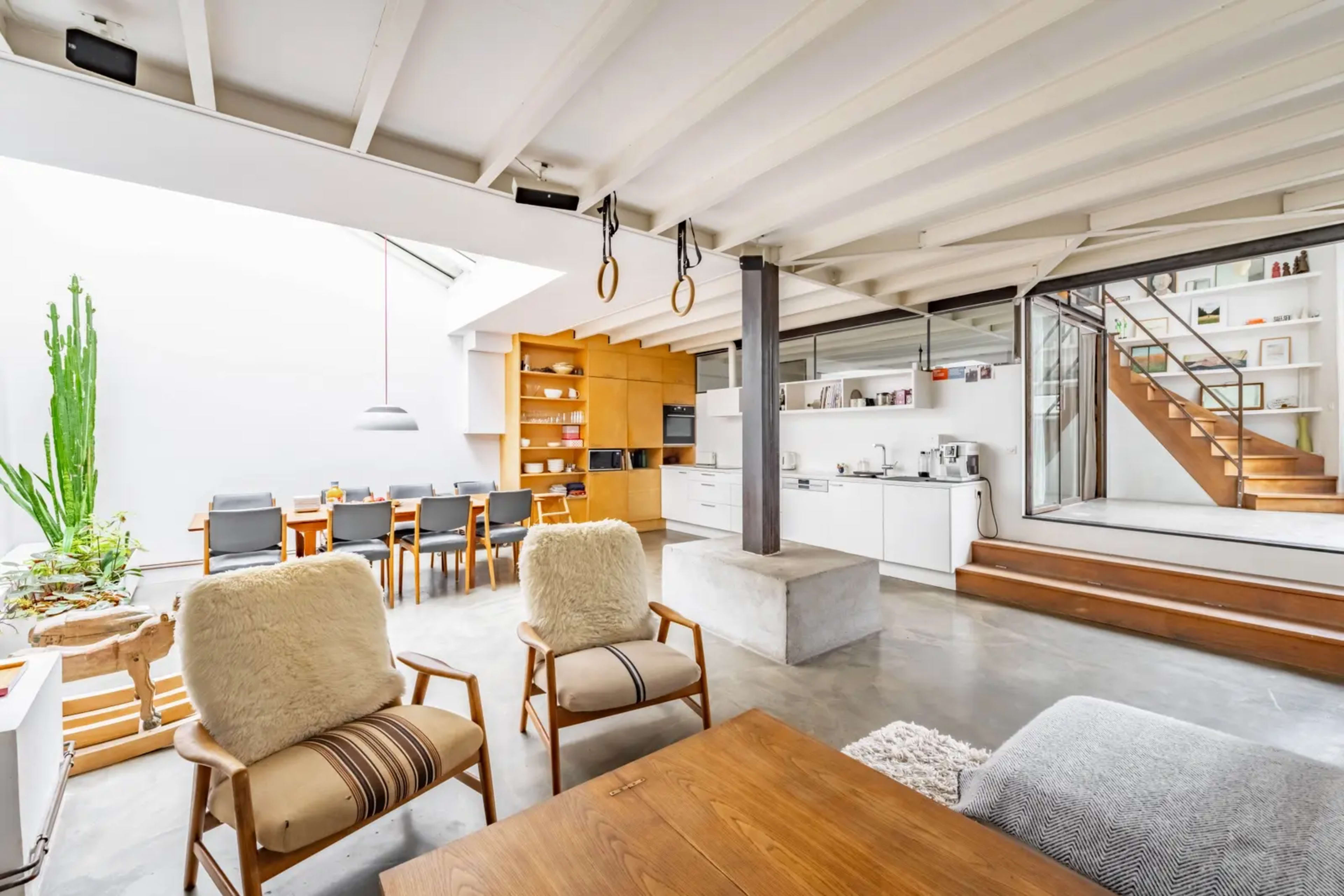 The image shows a modern, open-concept living space with a dining area, minimalist kitchen, and a staircase, surrounded by large windows allowing natural light.