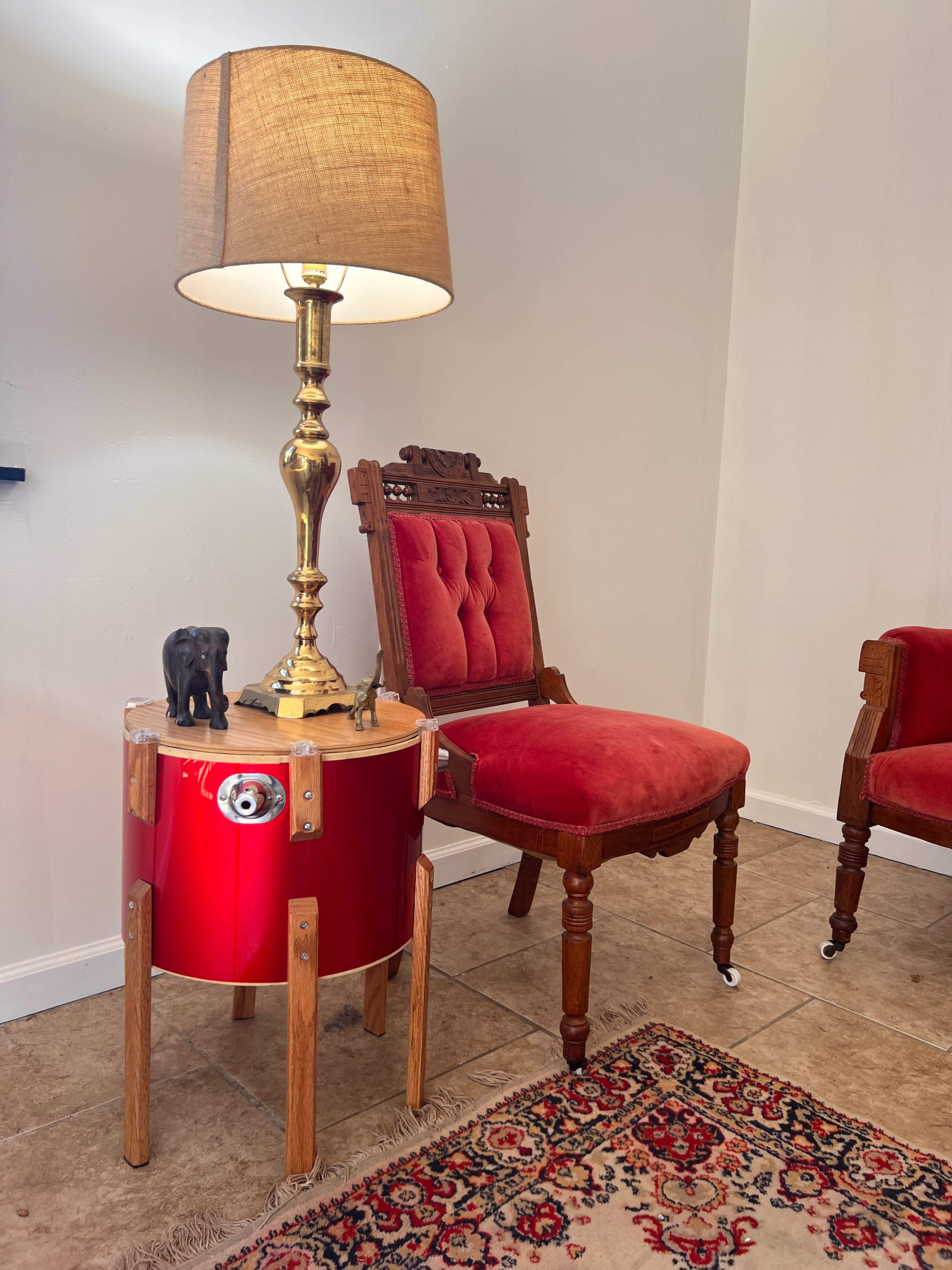 A red drum table with a brass lamp stands next to an ornate wooden chair upholstered in red fabric, set against a neutral wall.