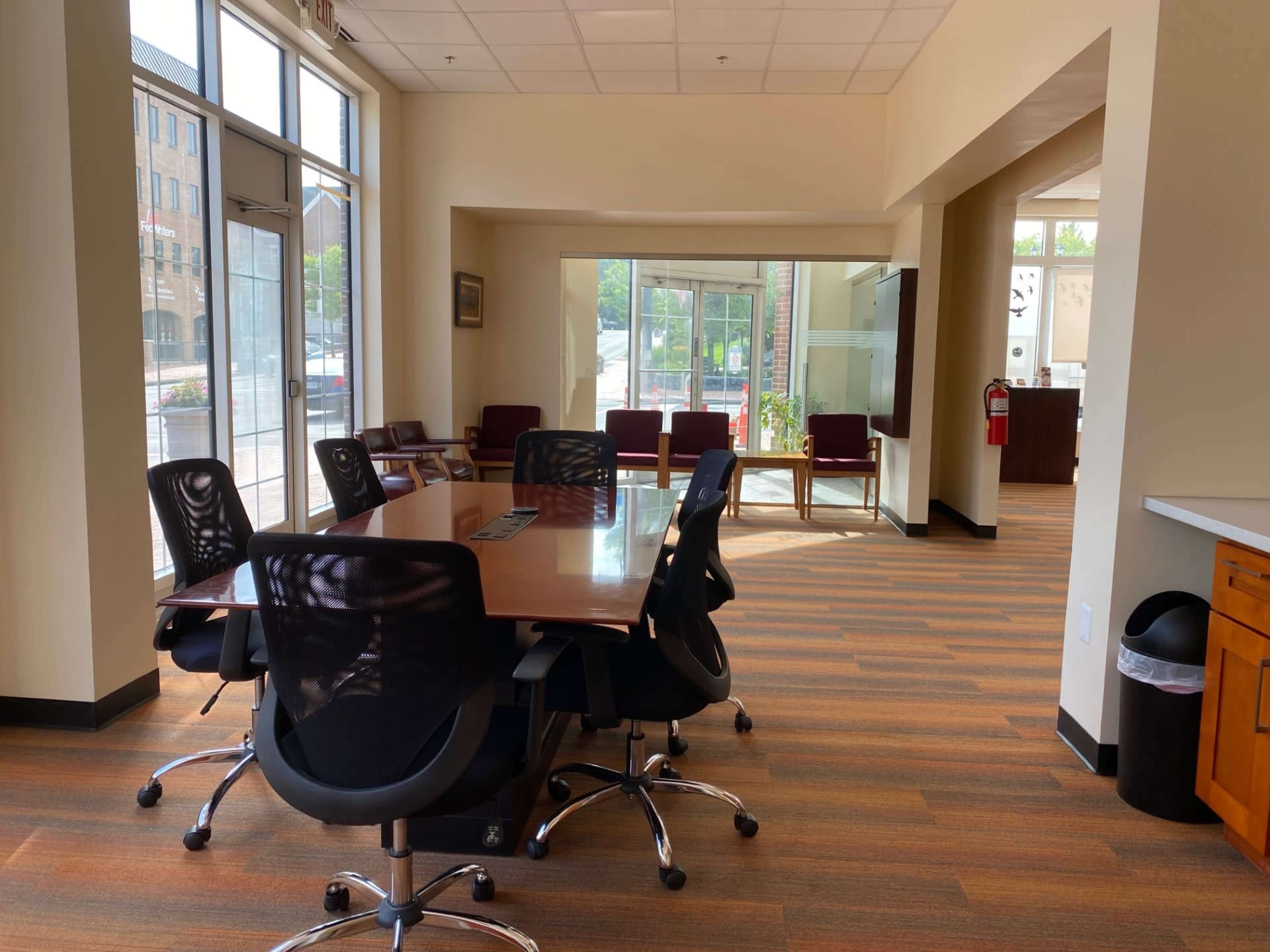 The image shows a modern office space featuring a large conference table surrounded by ergonomic chairs, with a view of seating and large windows in the background.