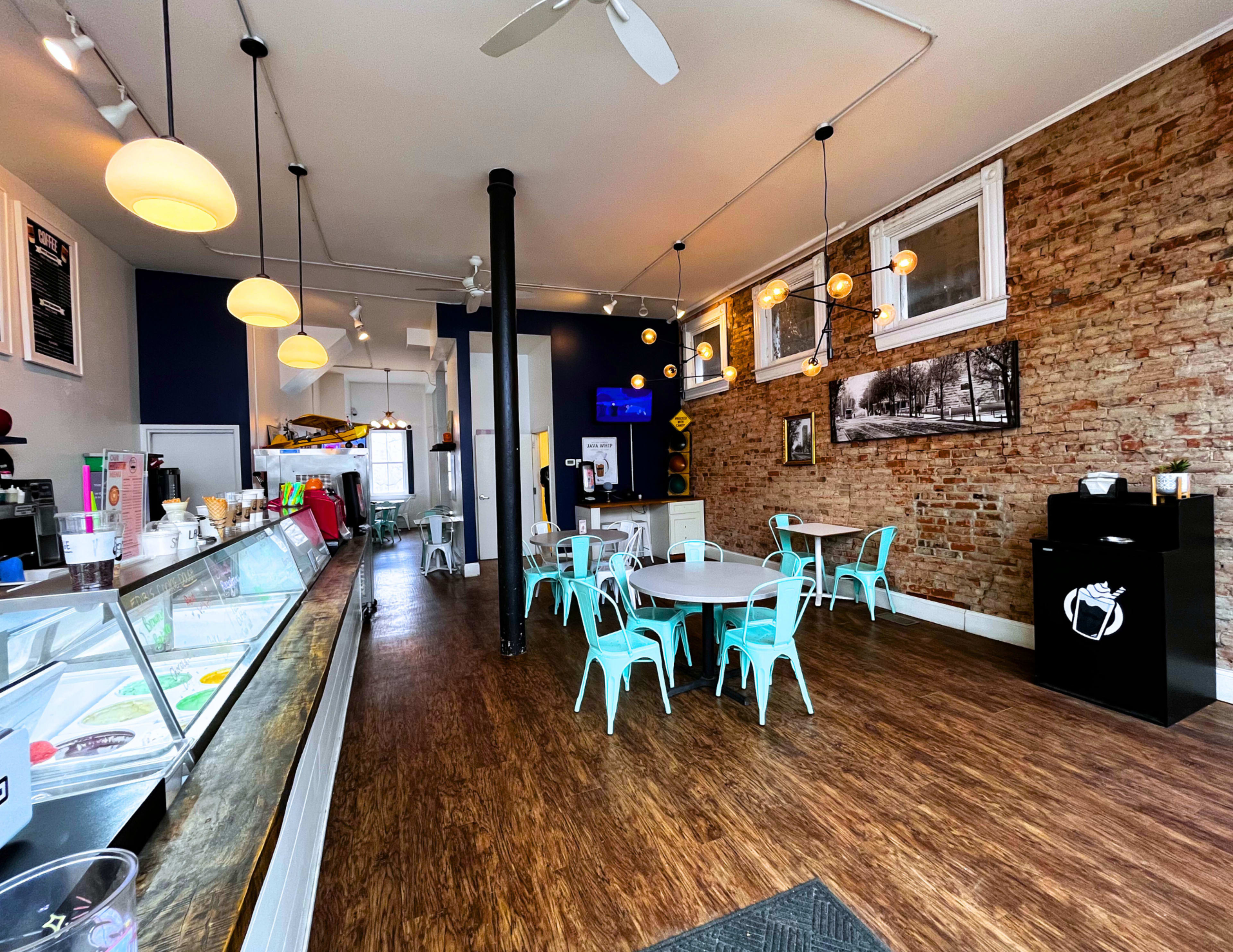 The image shows a small ice cream shop with a modern interior featuring turquoise chairs, a service counter displaying ice cream, and a brick wall backdrop.